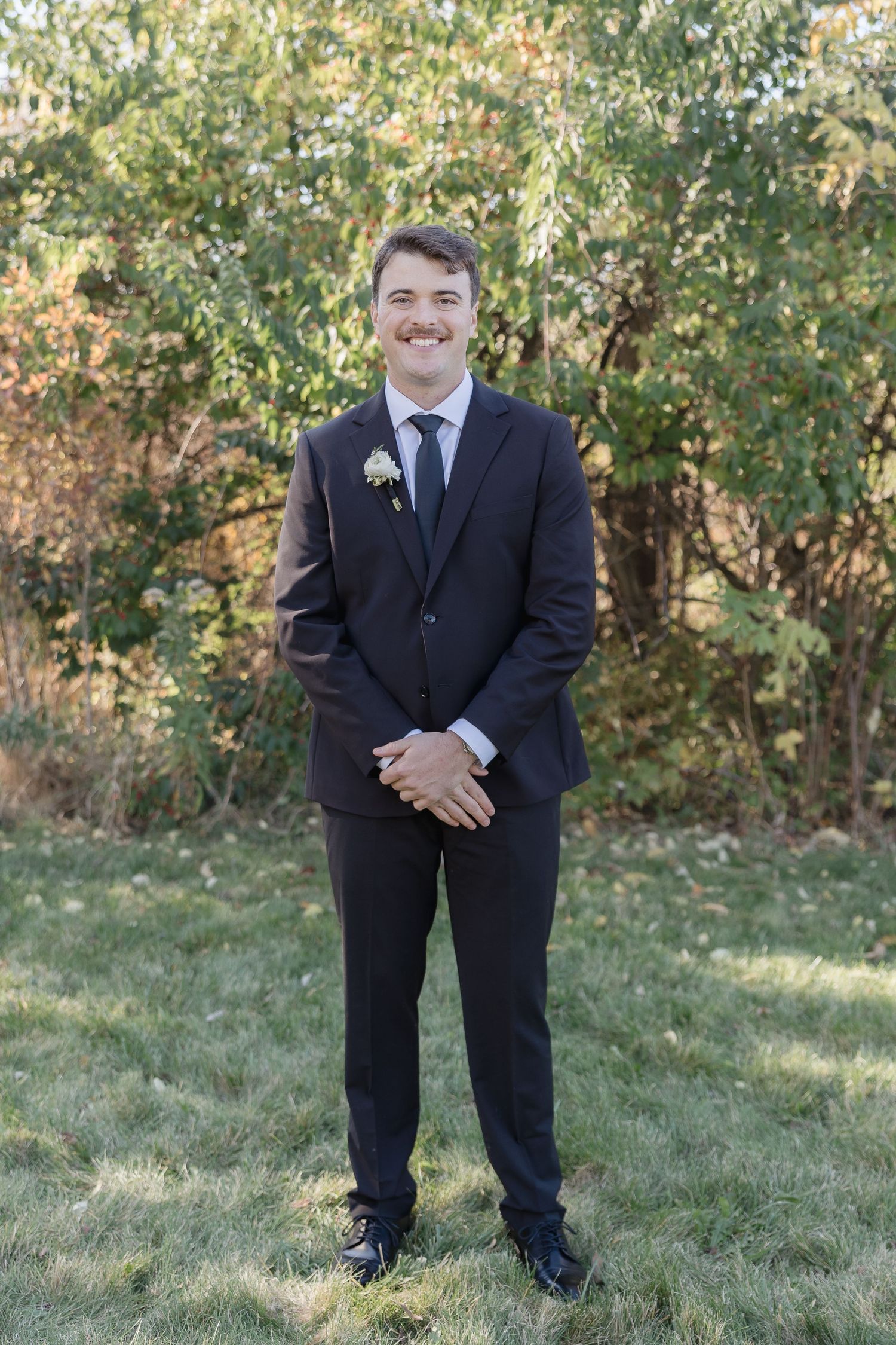 Someone wearing a dark suit and boutonniere stands on grass with autumn trees in the background for a formal occasion.