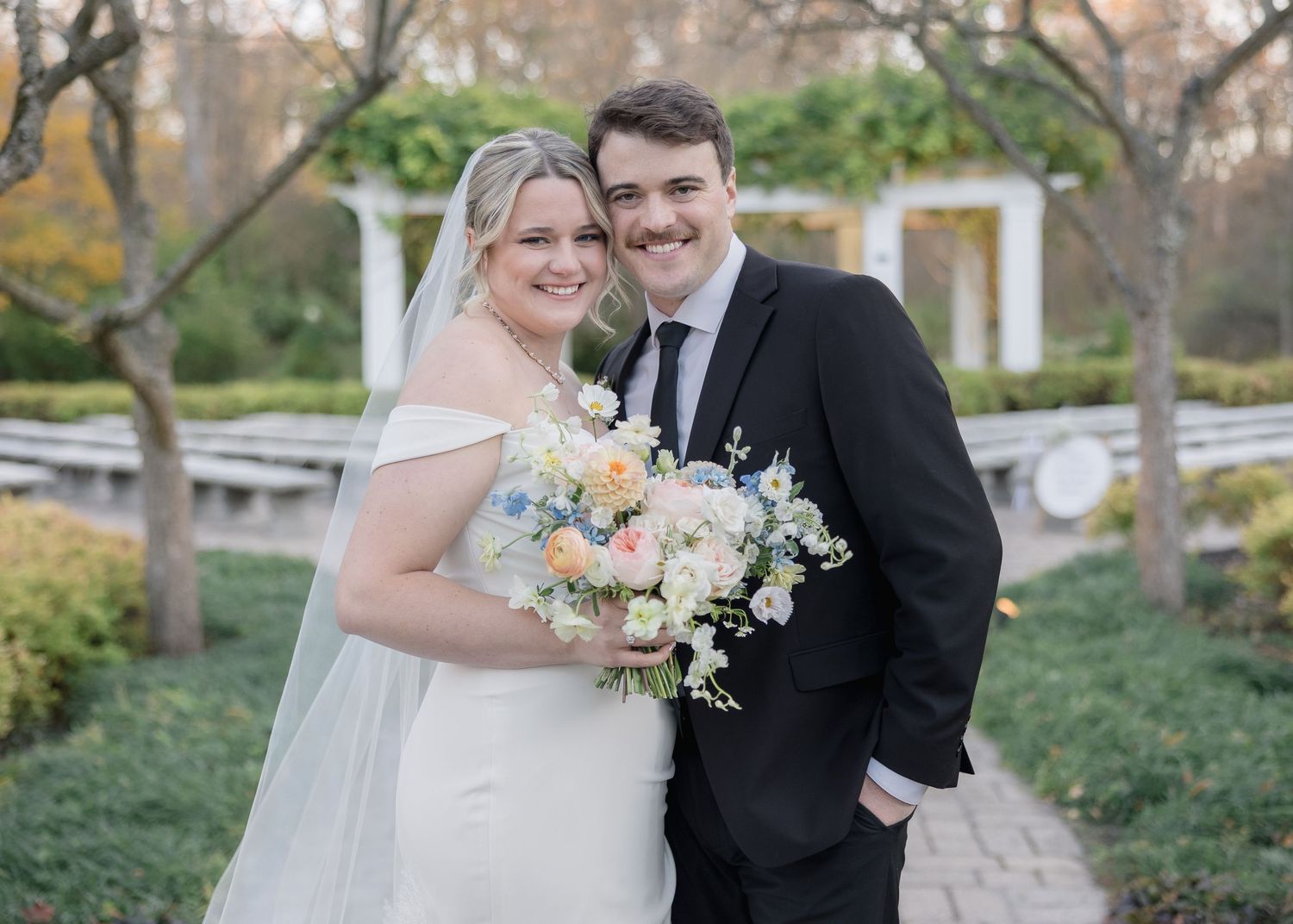 Elegant wedding portrait with bride in white dress holding pastel bouquet next to groom in black suit.