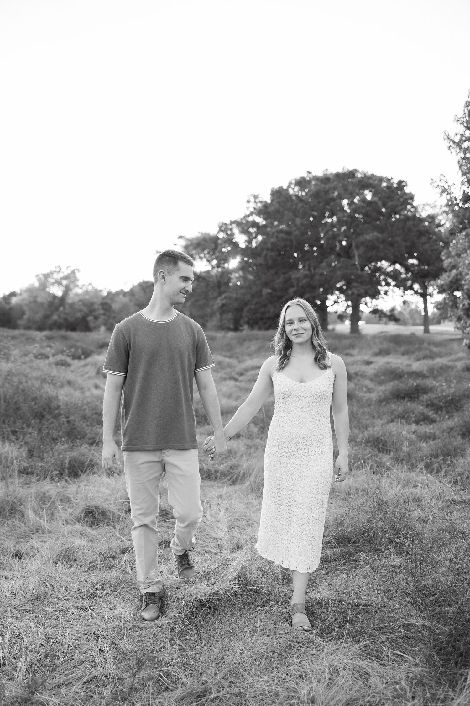 A couple holds hands while walking through a grassy field on a sunny day, captured in black and white.