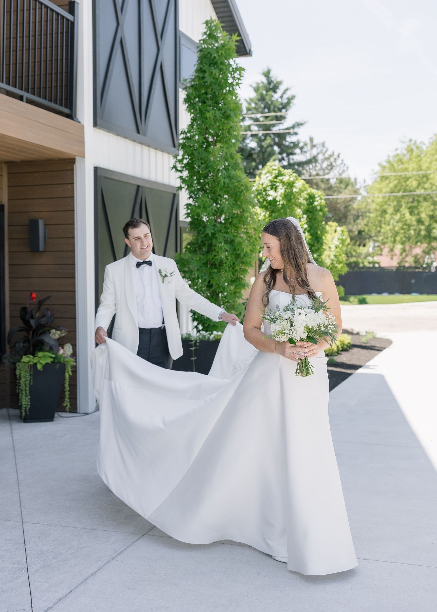 Groom in white tuxedo holds bride's flowing white gown as she admires her bouquet outside a modern venue.