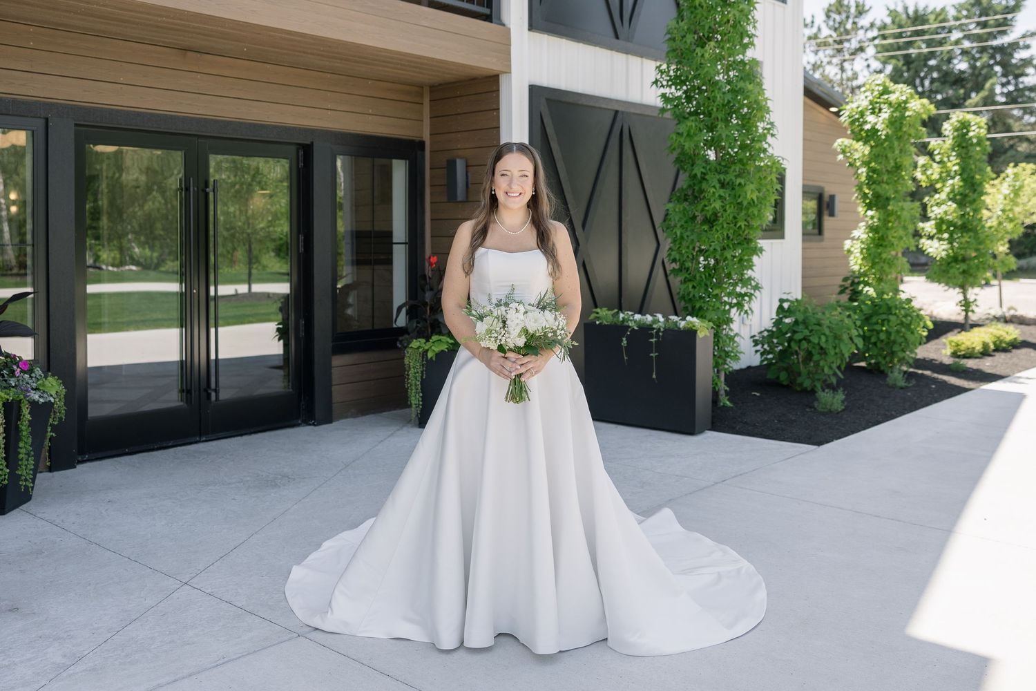 Bride in a white ball gown holding a bouquet stands outside a modern venue surrounded by lush greenery.