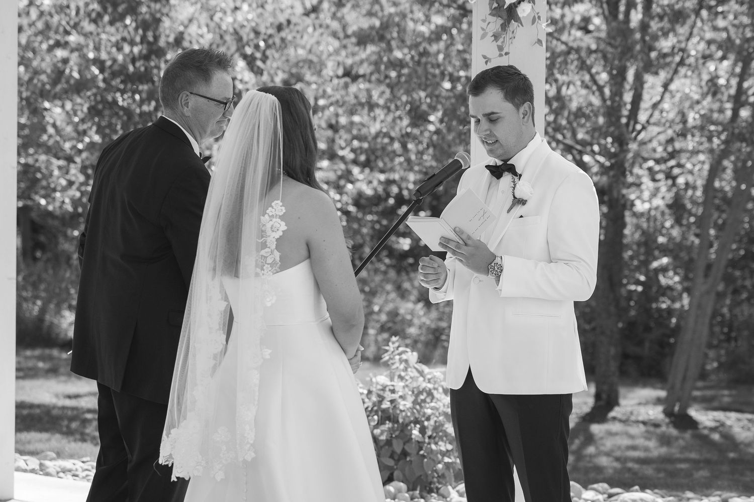 Black and white photo of a groom reading wedding vows to his bride during an outdoor ceremony with an officiant present.