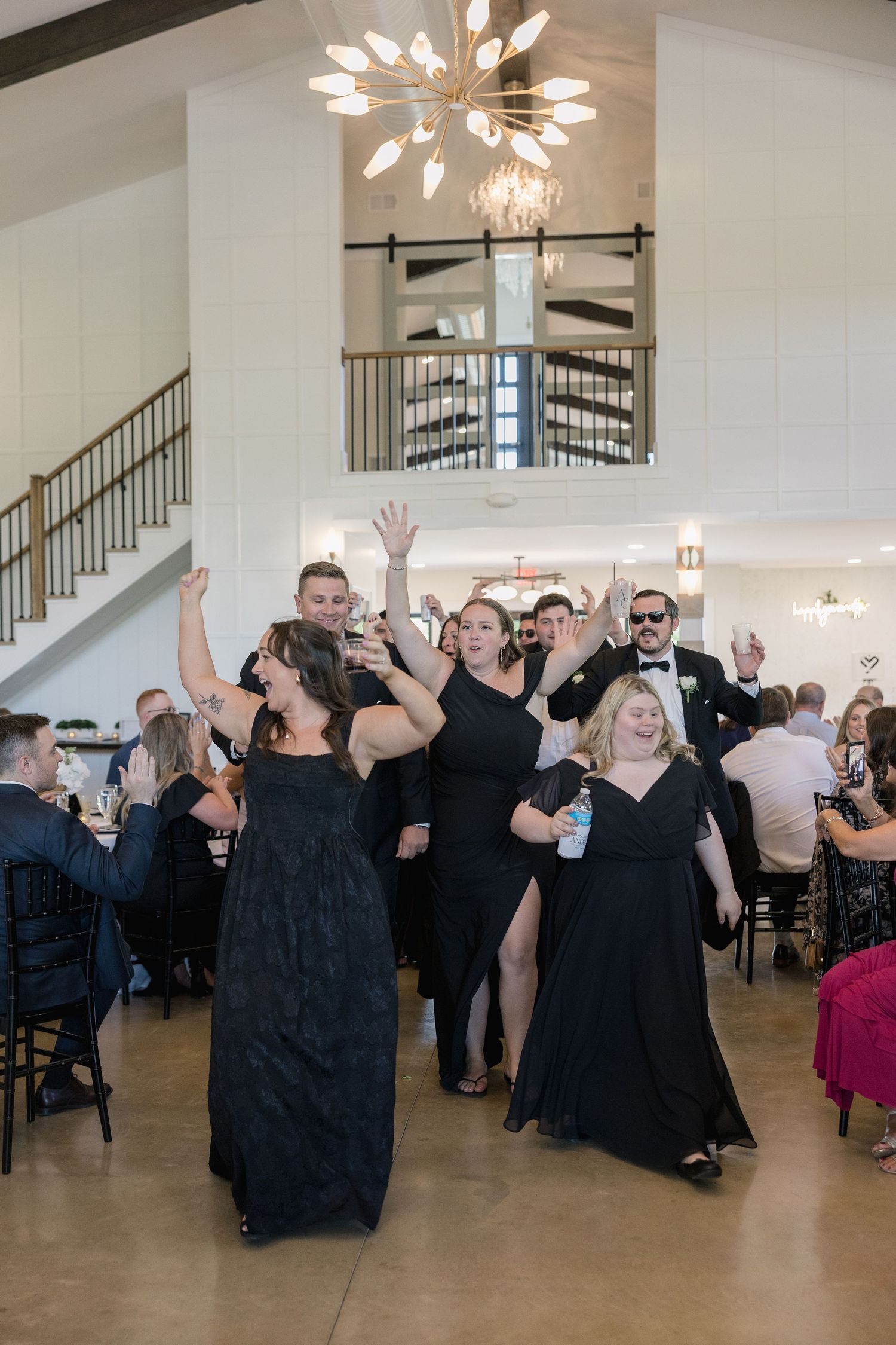 Wedding guests in black formal attire dancing and celebrating joyfully in an elegant white reception venue.