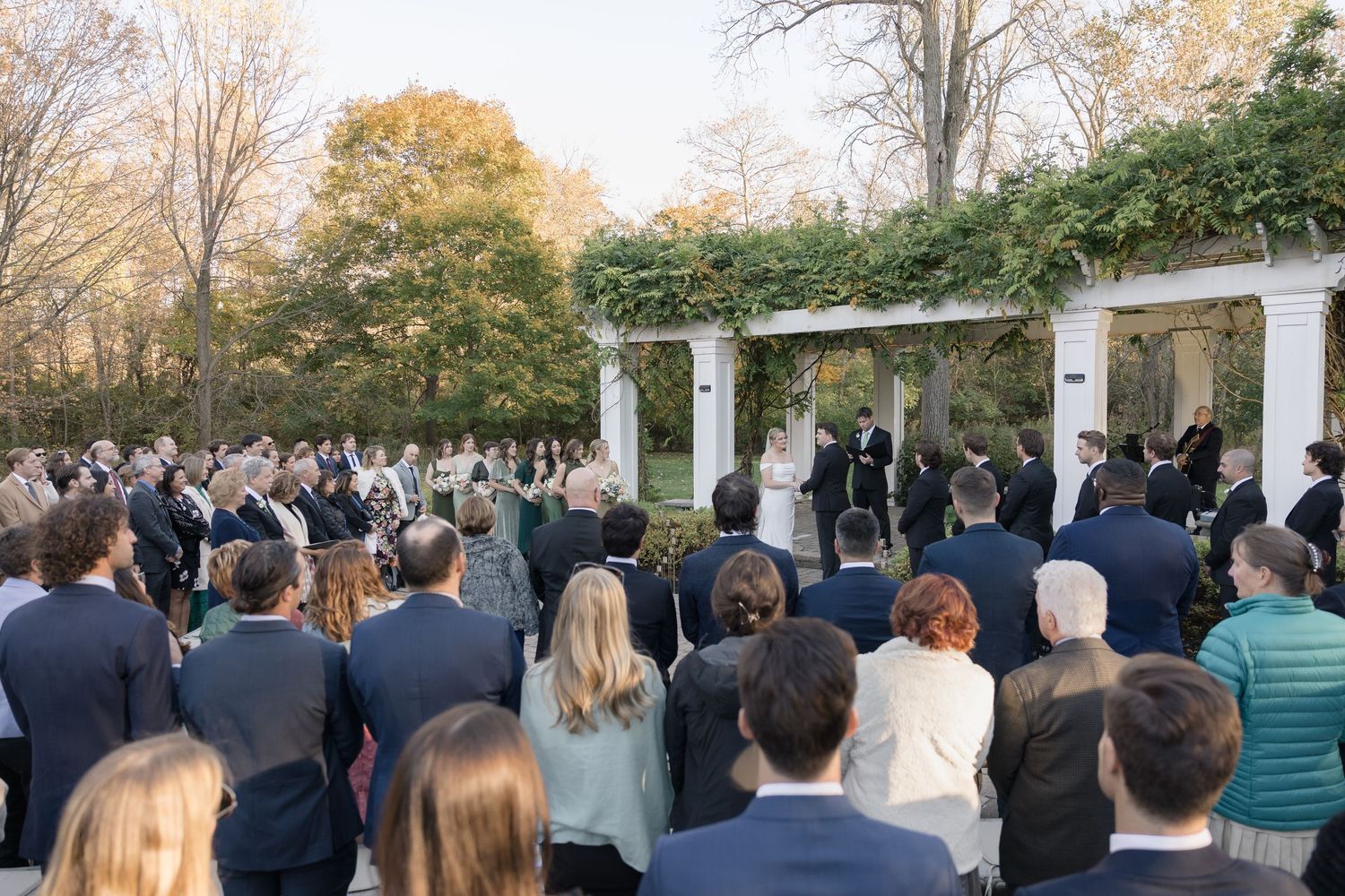 Large crowd of wedding guests watches outdoor ceremony under white columns with autumn foliage.