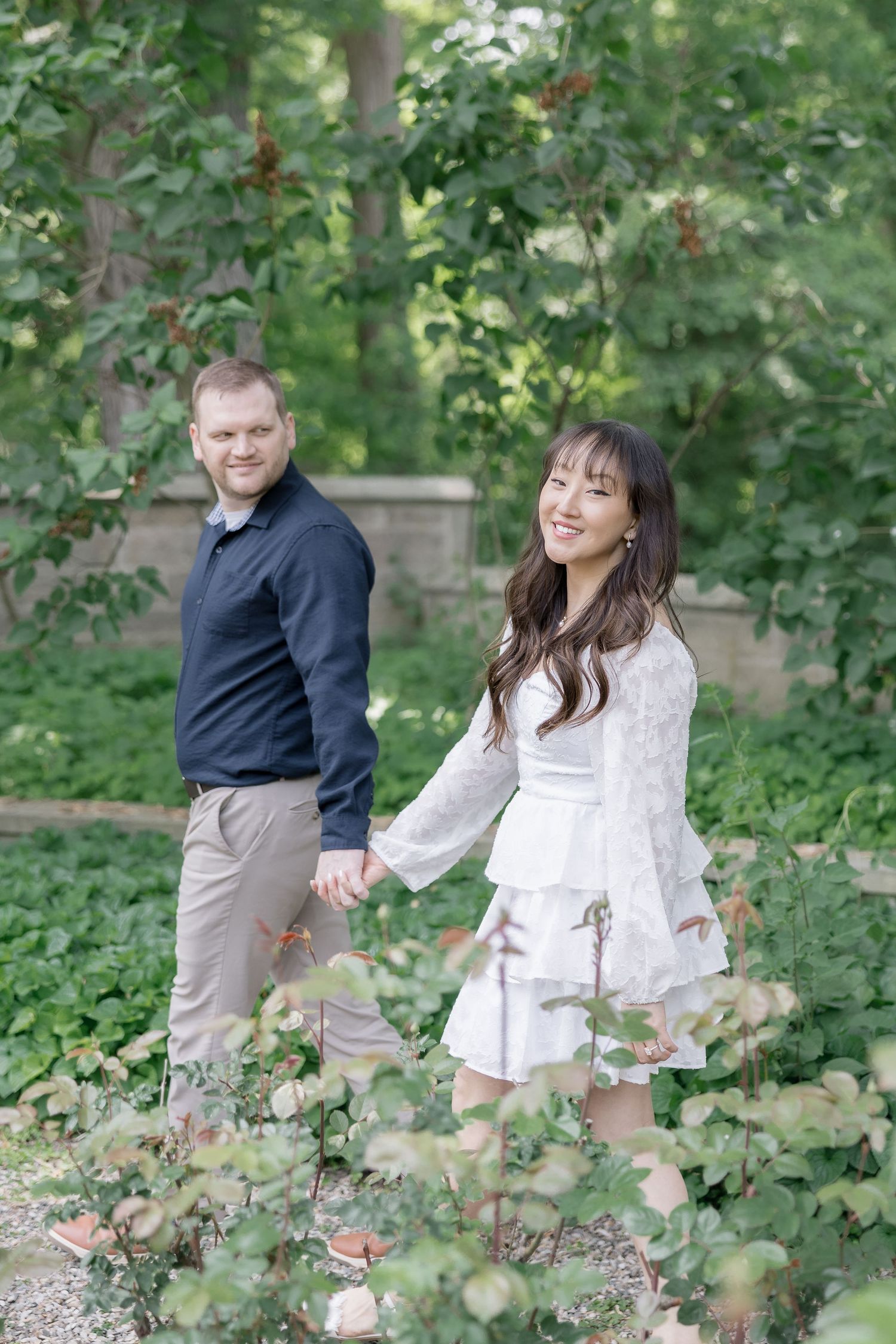 Couple holding hands while walking through a lush garden path with stone wall backdrop.