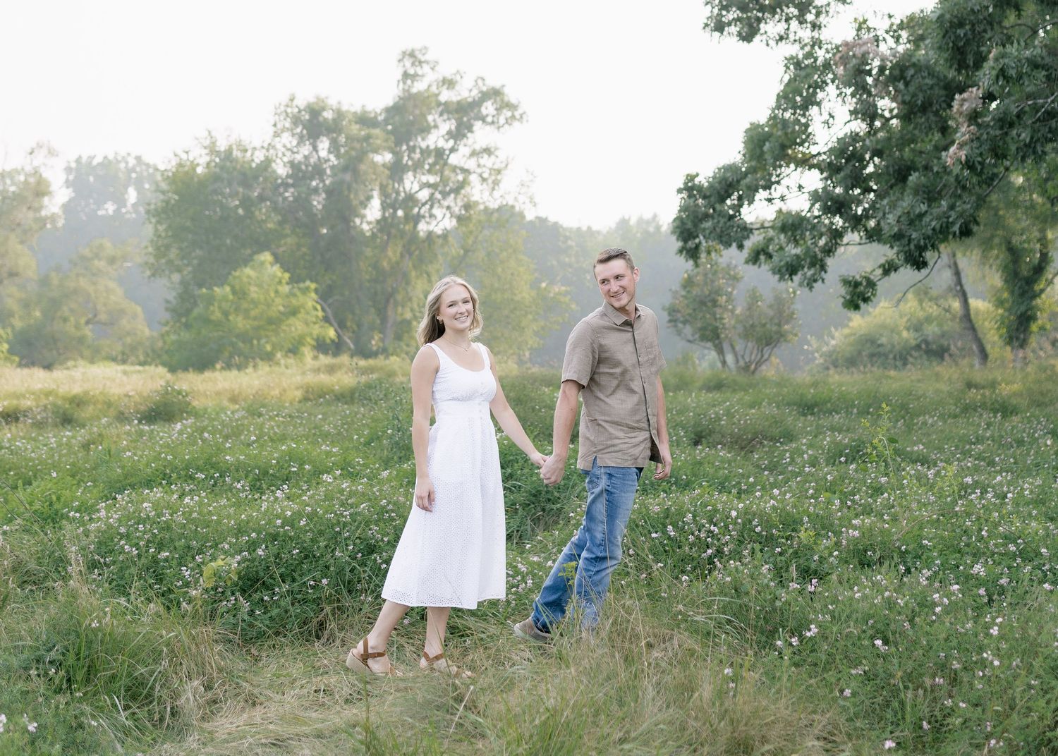 A couple walks hand in hand through a lush green meadow on a misty summer evening.