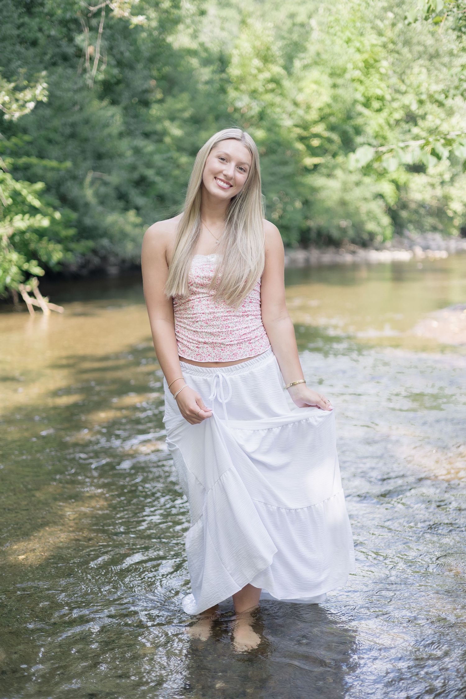 A summer portrait taken at a creek with someone wearing a pink top and flowing white skirt standing in shallow water.