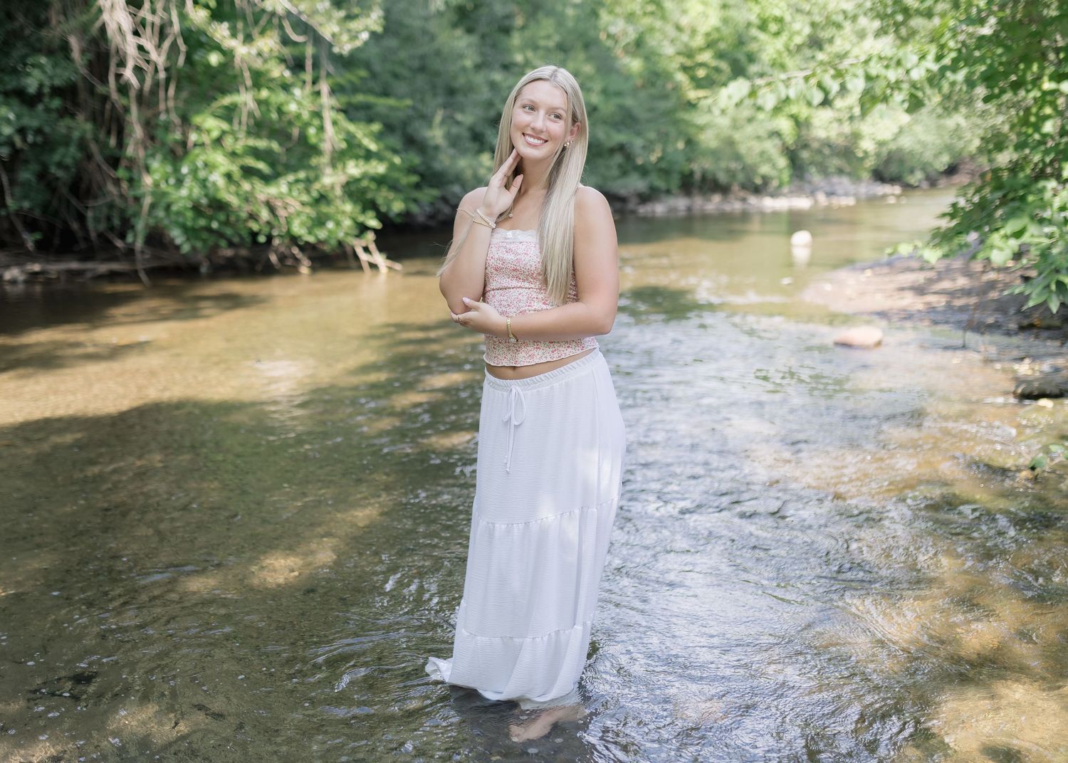 A person in a white dress stands in a flowing creek surrounded by lush green trees on a summer day.