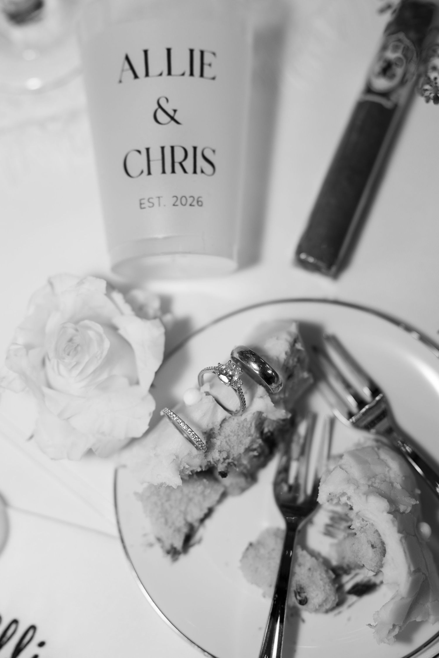 Black and white photo of wedding rings on a cake plate beside a rose and Allie & Chris EST. 2026 sign.