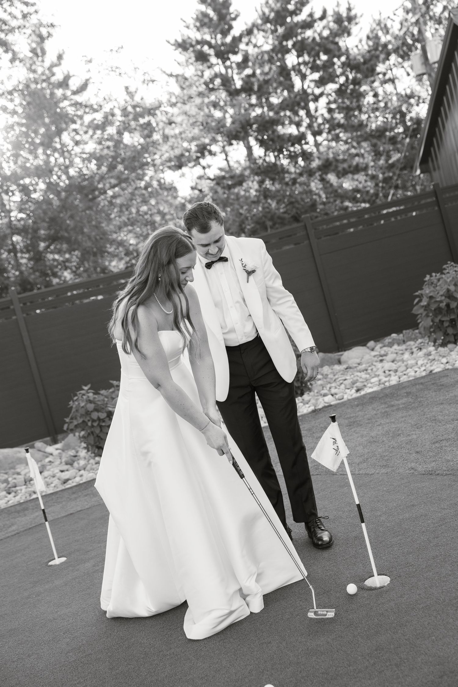 Black and white photo of groom teaching bride to putt golf on wedding day outdoor putting green.