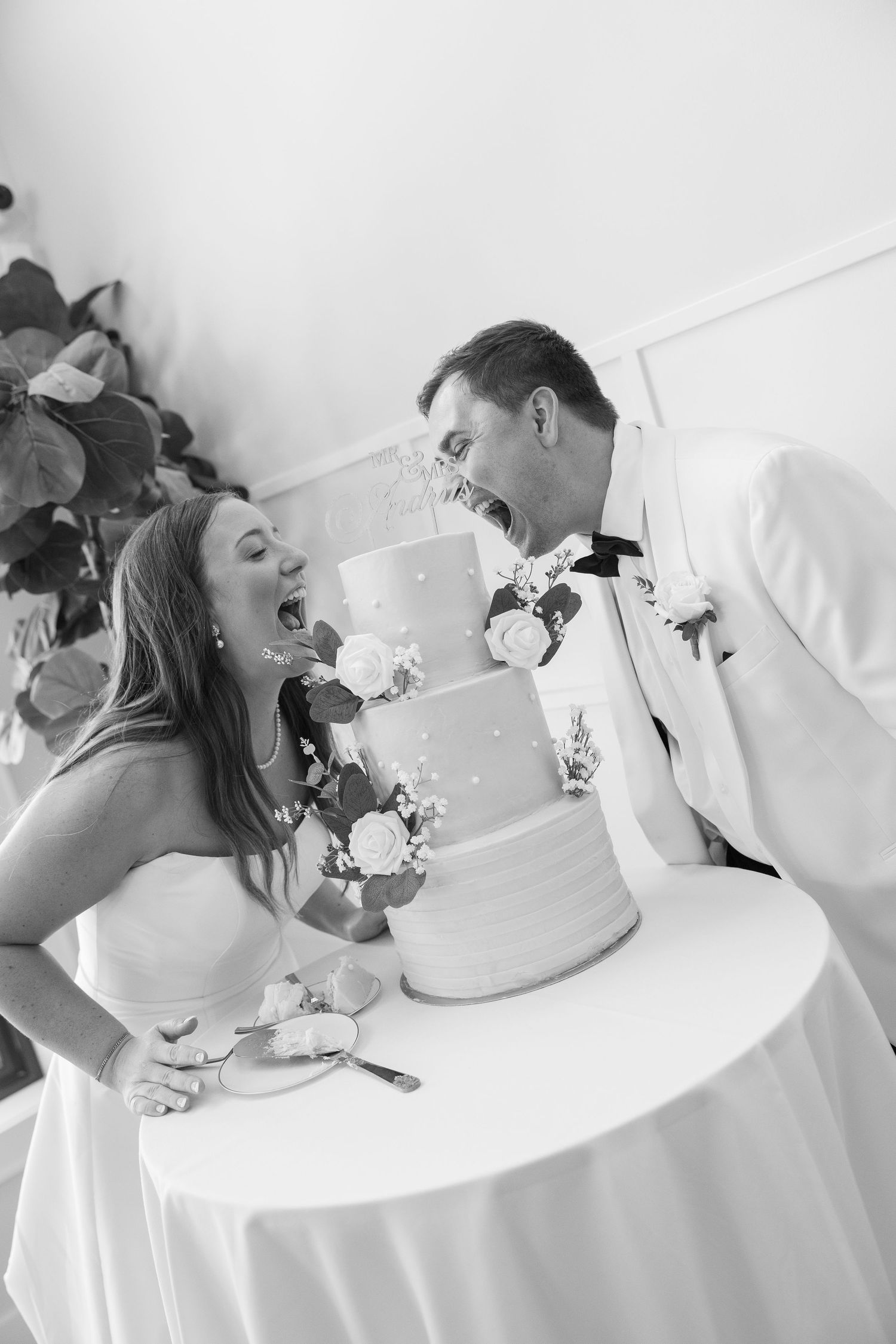 Bride and groom joyfully eating wedding cake together in black and white photo at their reception.