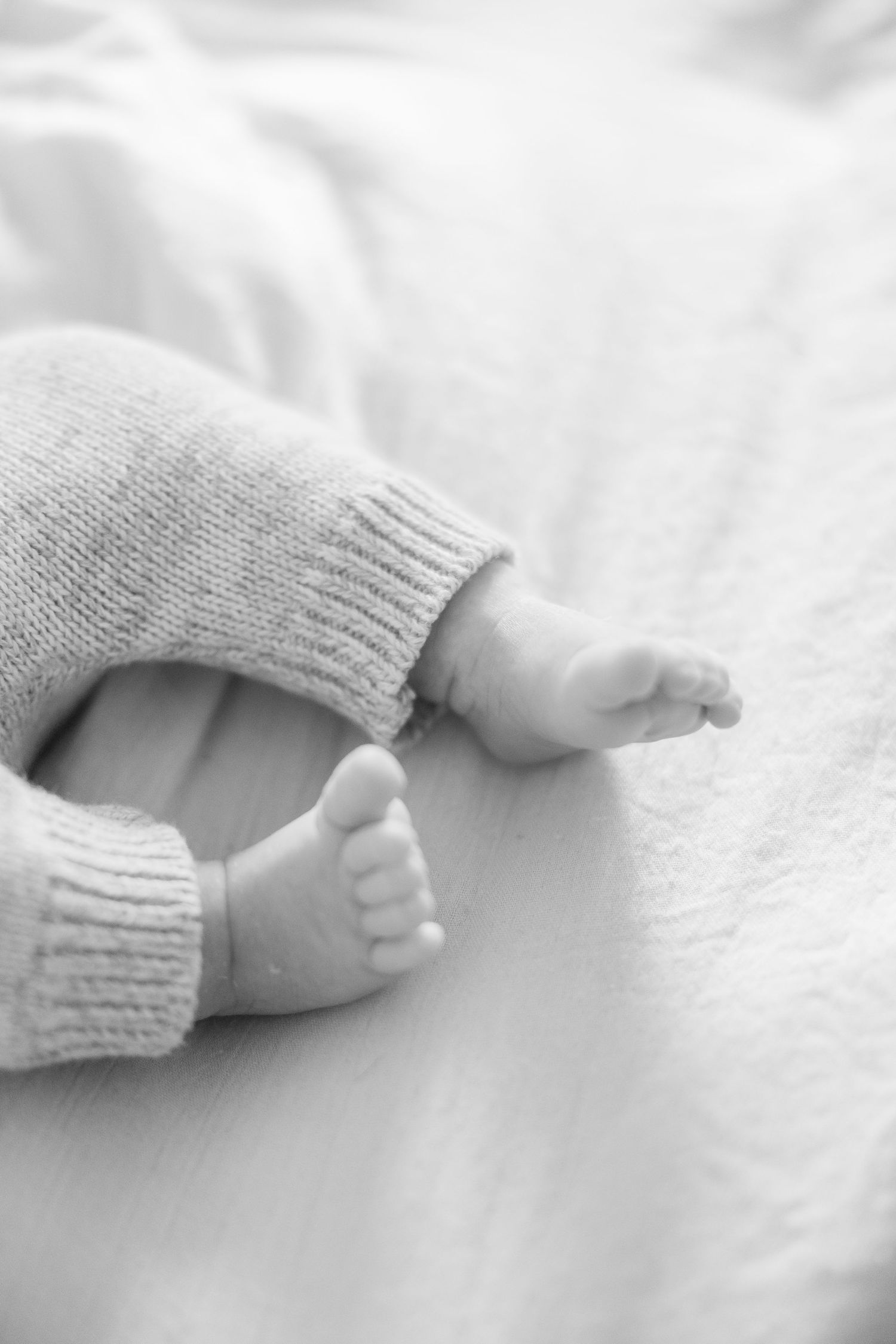Baby feet in gray knit booties rest on white bedding in soft natural light.