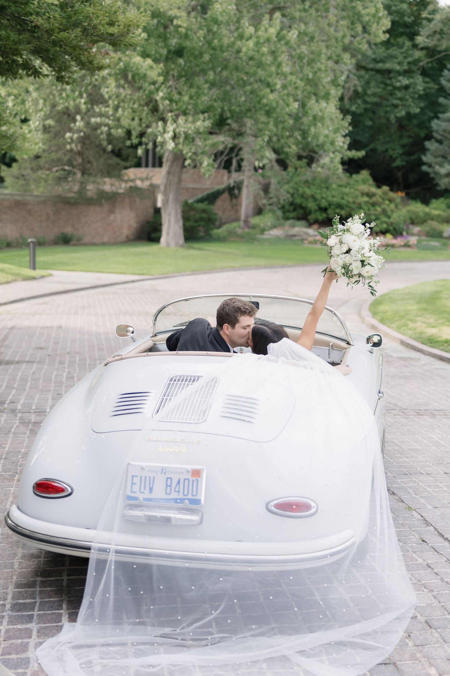 Newlywed couple kissing in a vintage white Porsche convertible, bride holding bouquet with veil flowing, on a brick driveway.