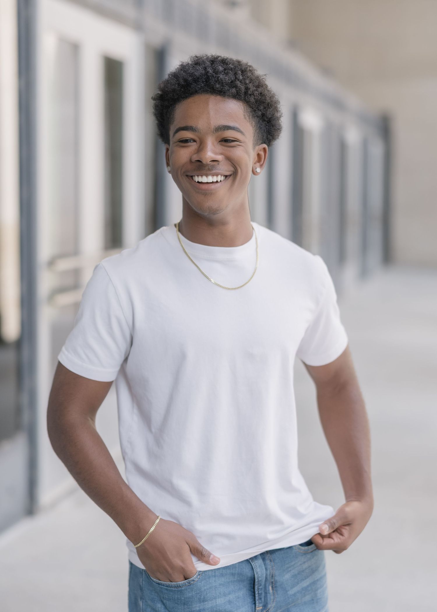 A person wearing a white t-shirt and jeans stands in front of a light-colored wall with a bright smile.