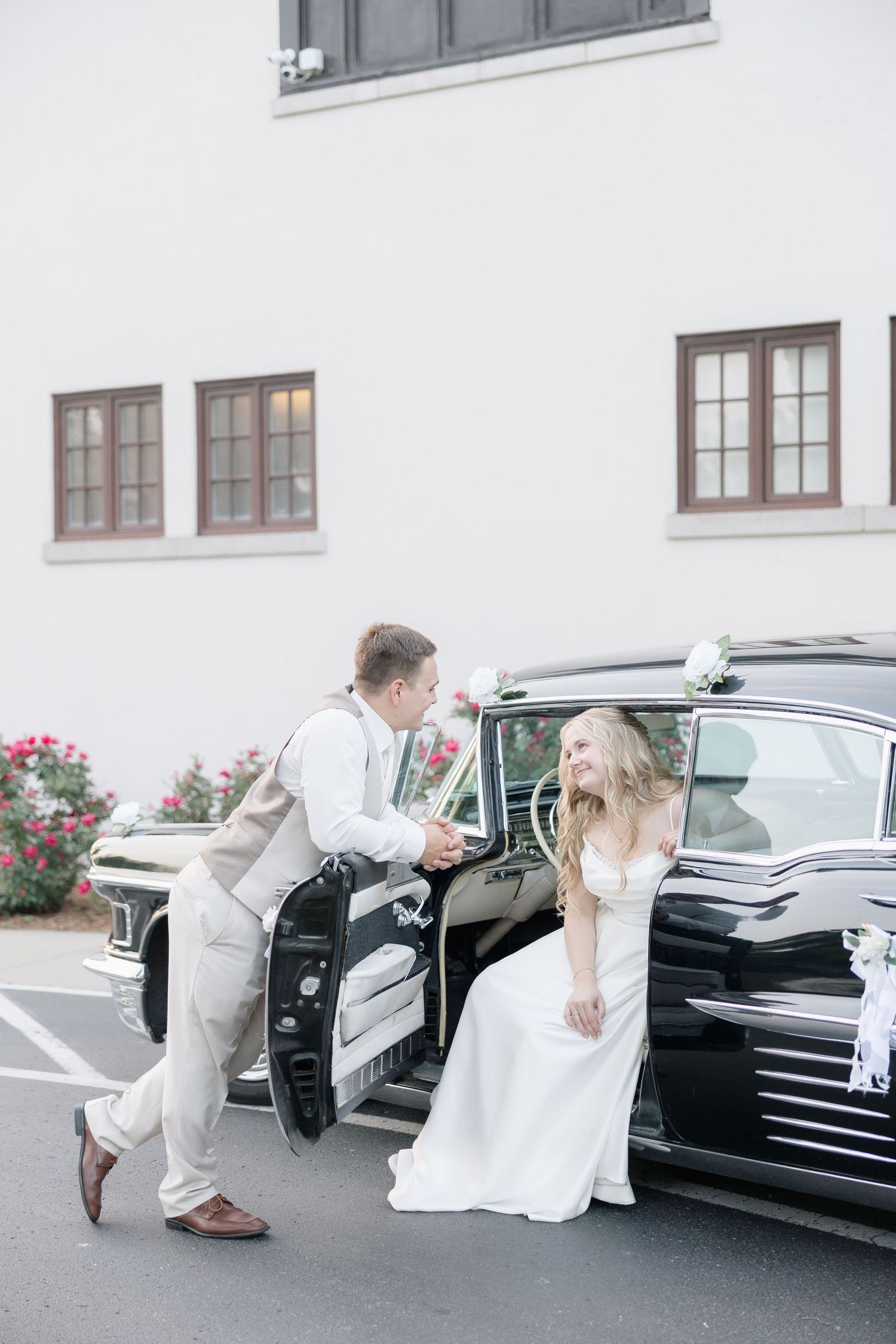 Wedding couple interacting playfully near vintage car in formal attire.