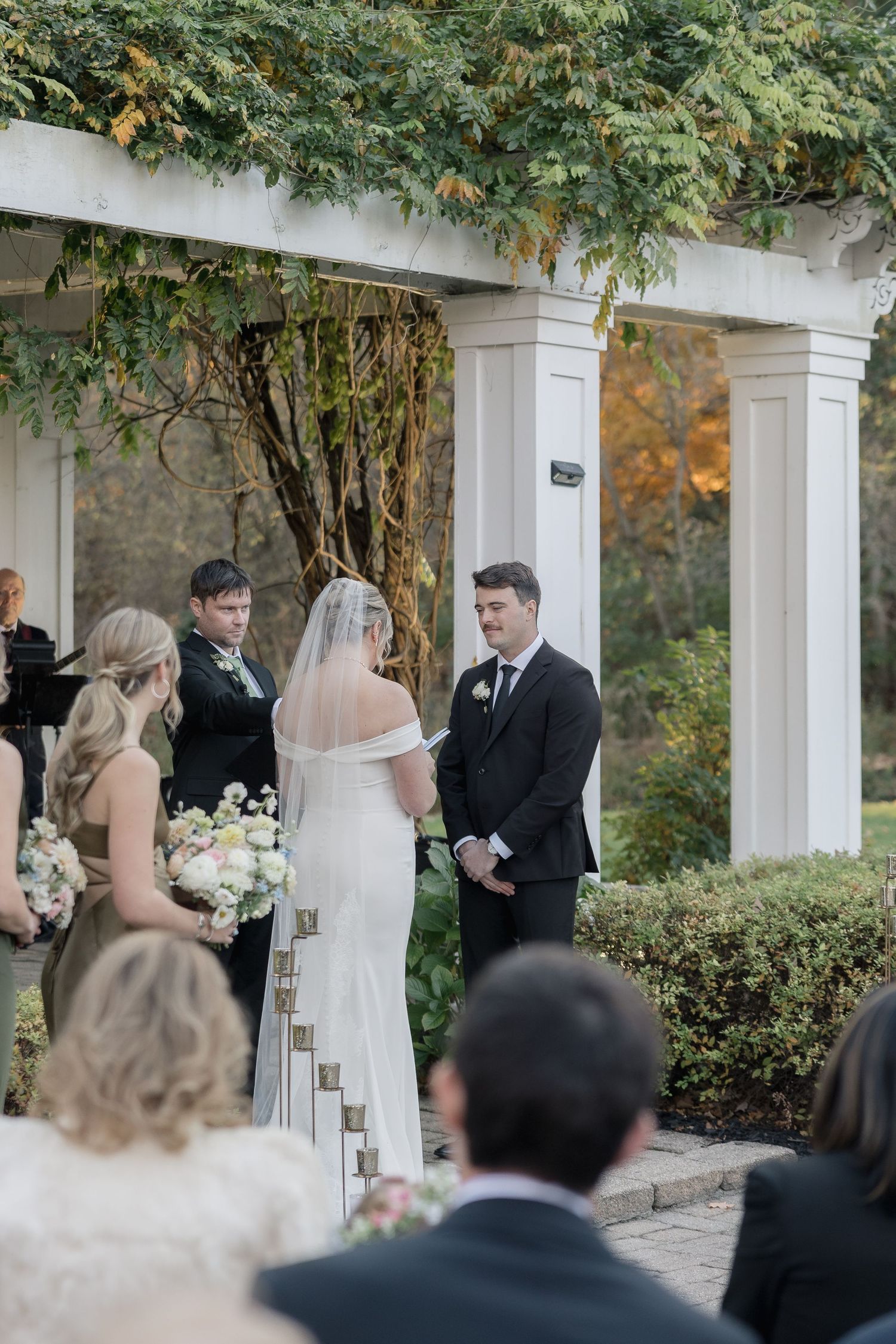 Outdoor wedding ceremony with guests watching under white columned structure adorned with greenery.