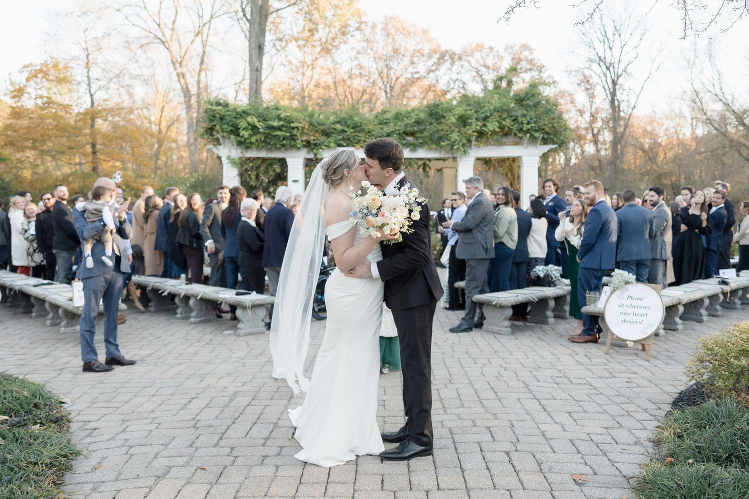 A wedding ceremony takes place outdoors on a stone patio with guests seated on wooden benches under an autumn sky.