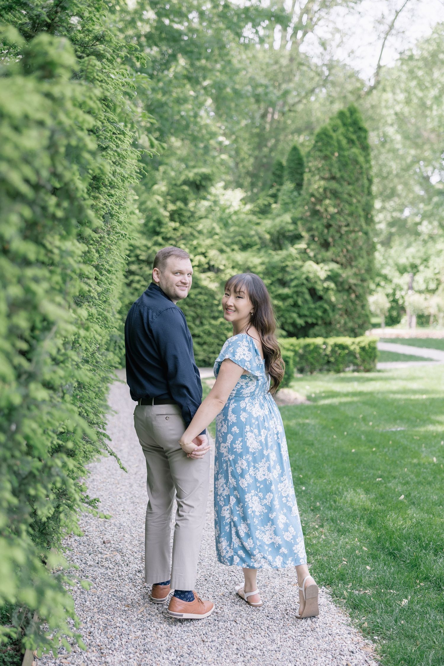 A couple walks and poses together on a gravel path surrounded by green foliage in a garden setting.