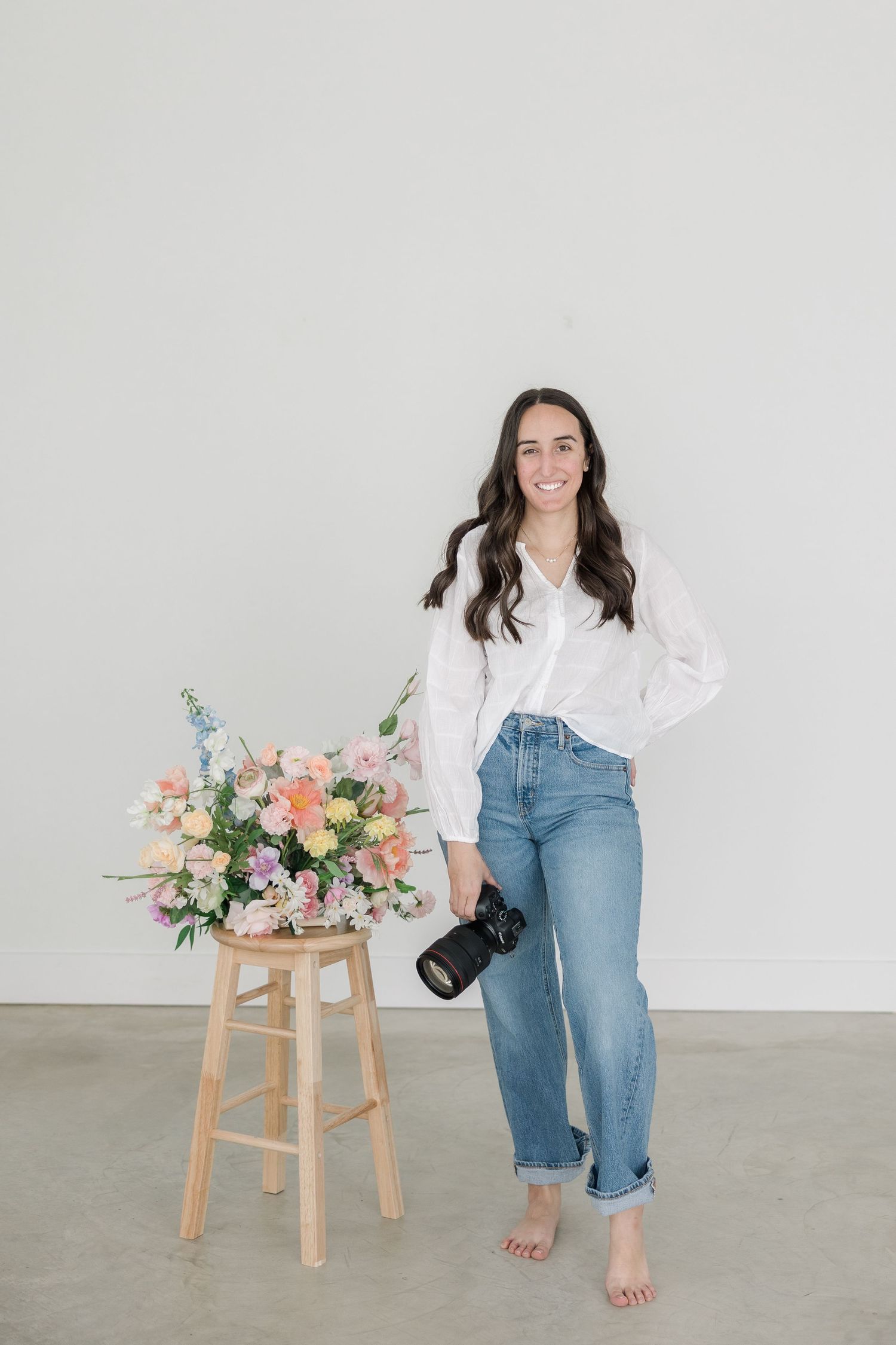 A young woman with long dark hair stands next to a wooden stool with a bouquet of colorful flowers on it, wearing a white blouse and jeans.