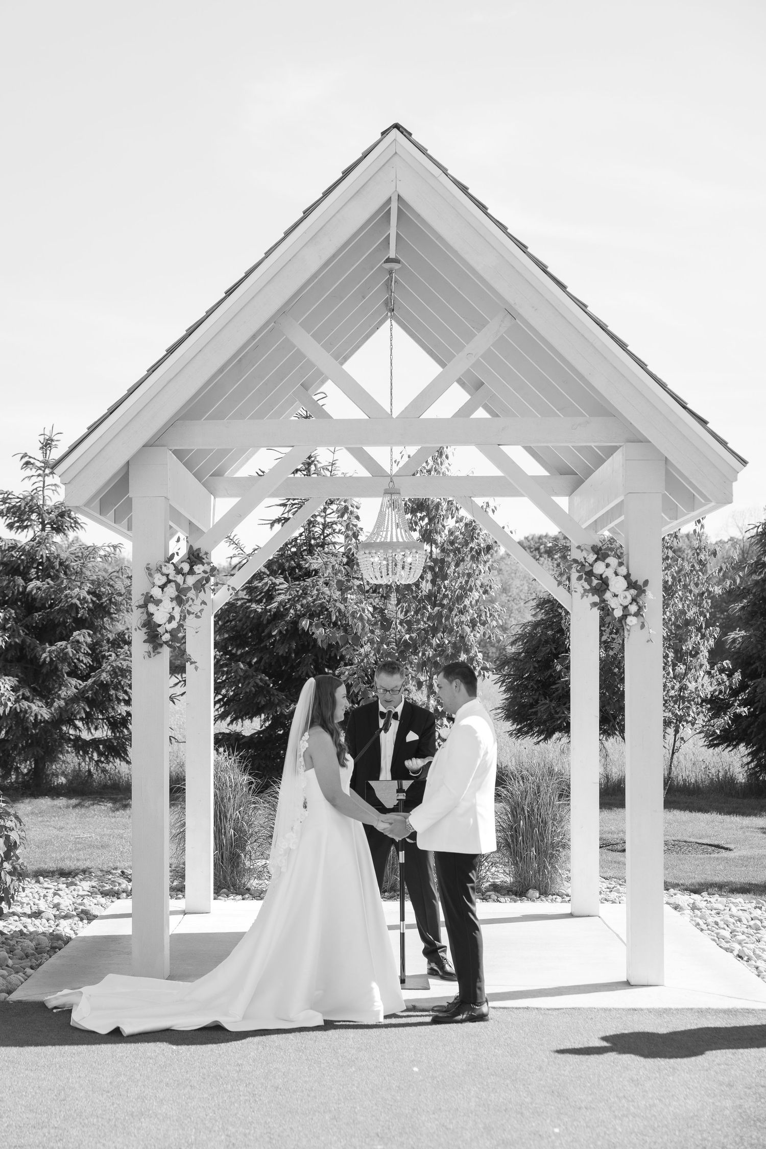 Black and white photo of a bride and groom exchanging vows under a white wooden gazebo decorated with flowers.