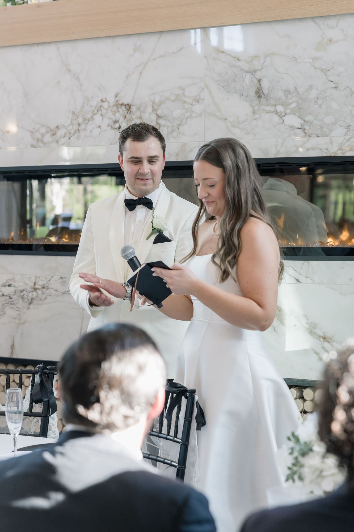 Bride and groom reading wedding vows together at reception, standing before marble fireplace in elegant venue.