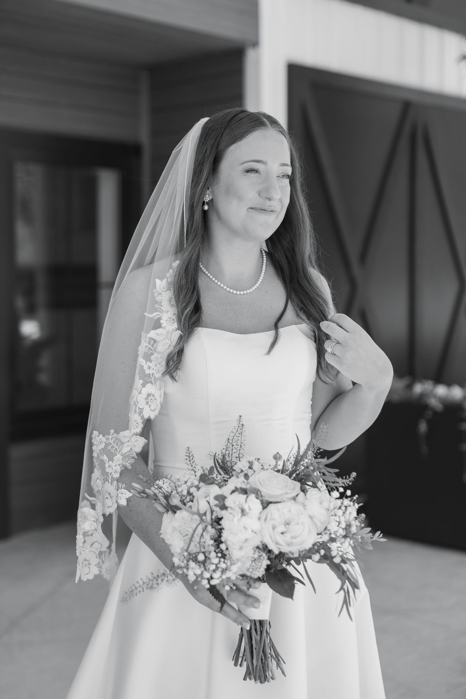 A bride in a white gown and lace veil holds a floral bouquet in a classic black and white portrait.