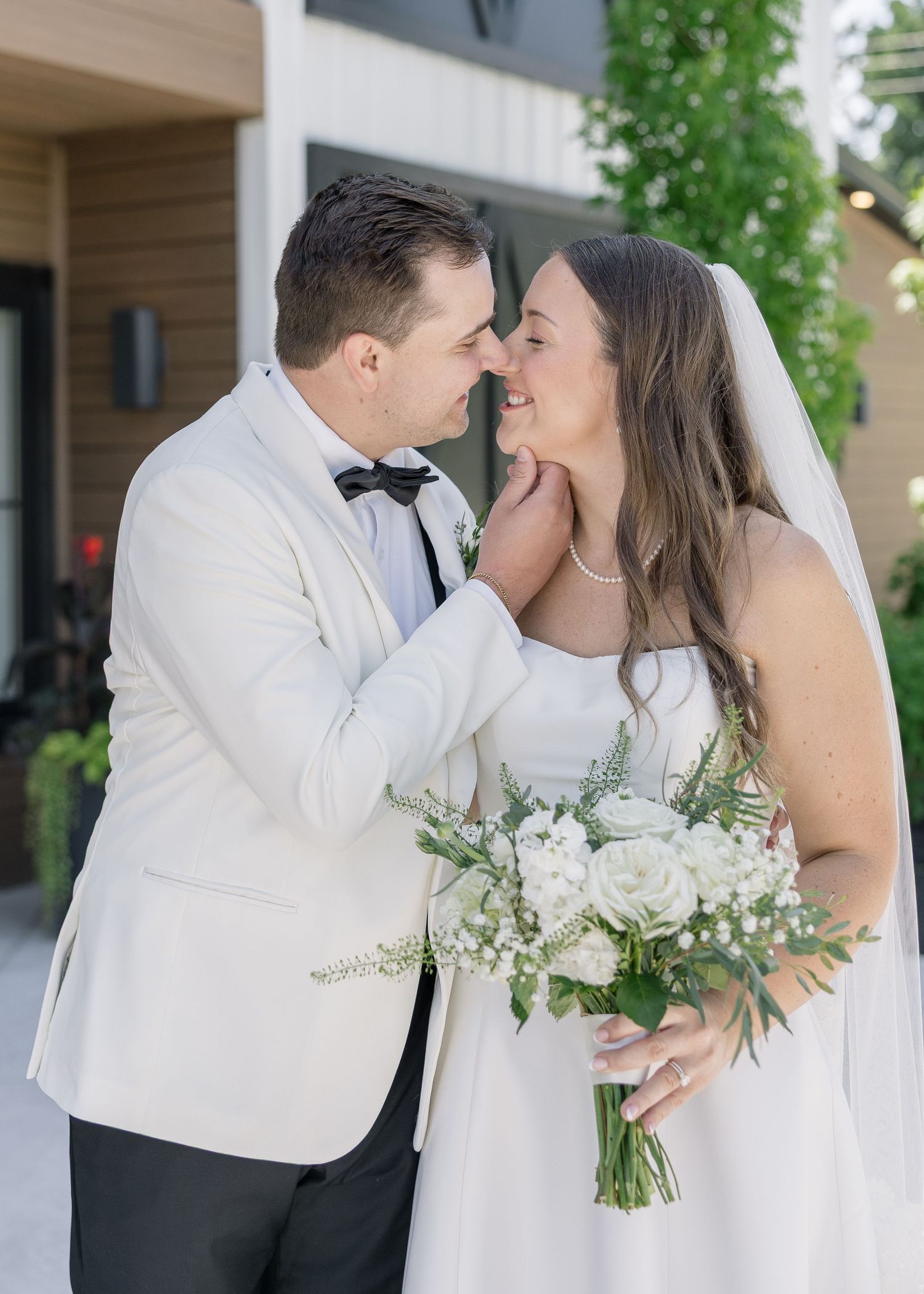 A bride and groom share a romantic moment, with the groom in a white tuxedo and bride holding a white floral bouquet.