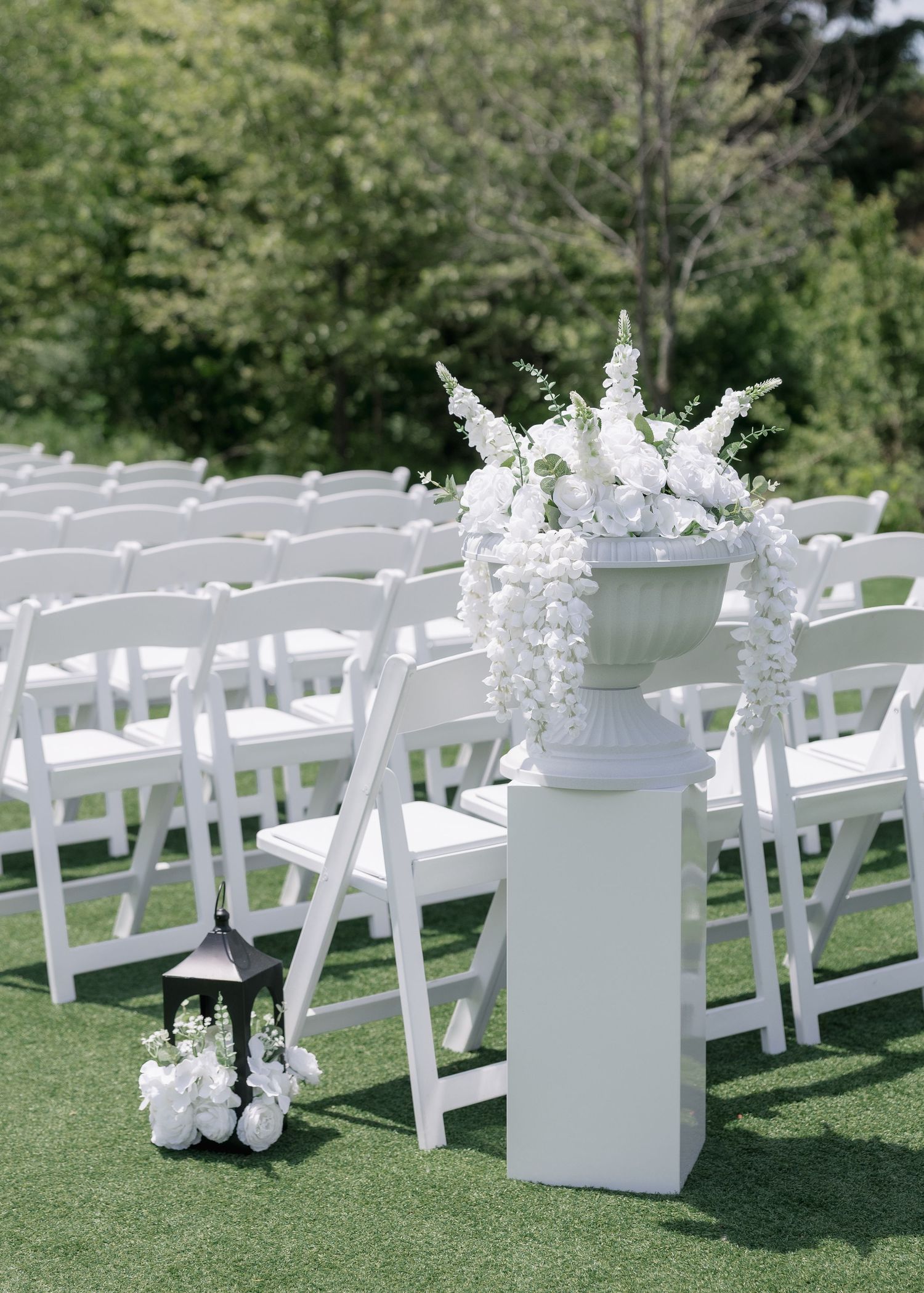 White folding chairs arranged on grass for an outdoor wedding ceremony with white floral arrangements on pedestals.