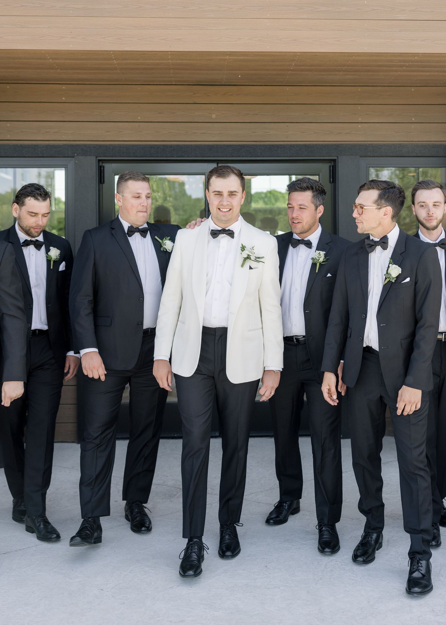Groom in white tuxedo jacket stands with groomsmen in black suits and bow ties outside modern building.