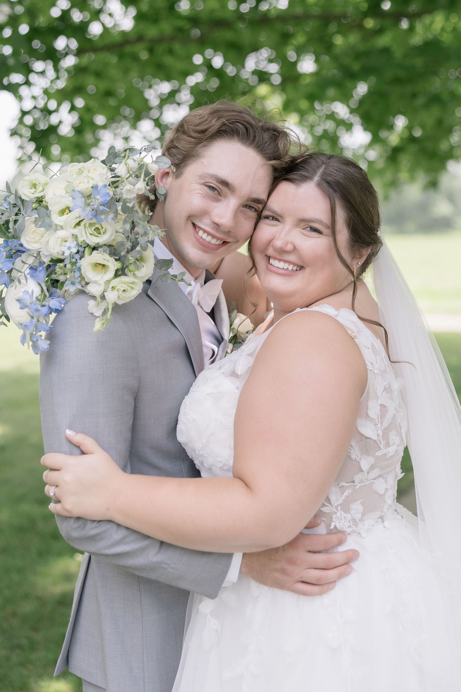 A joyful wedding couple embraces outdoors surrounded by lush greenery and white floral bouquets.