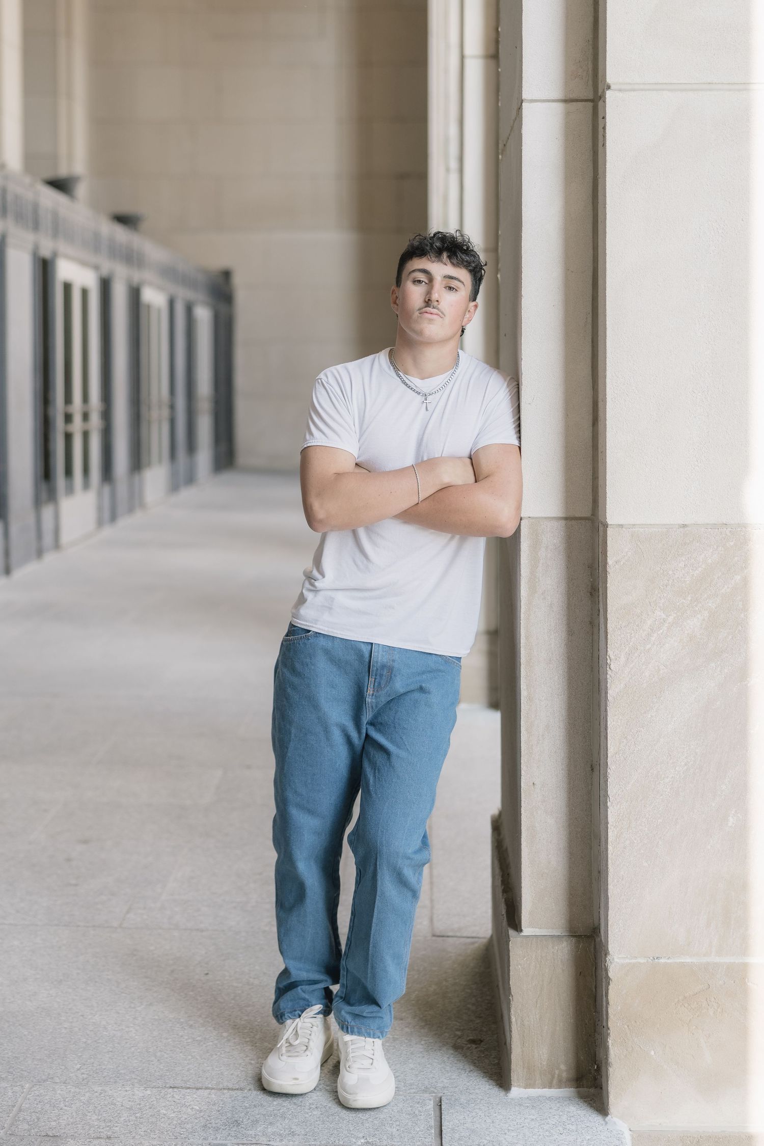 Person wearing white t-shirt and blue jeans poses casually against beige pillars in a modern corridor.