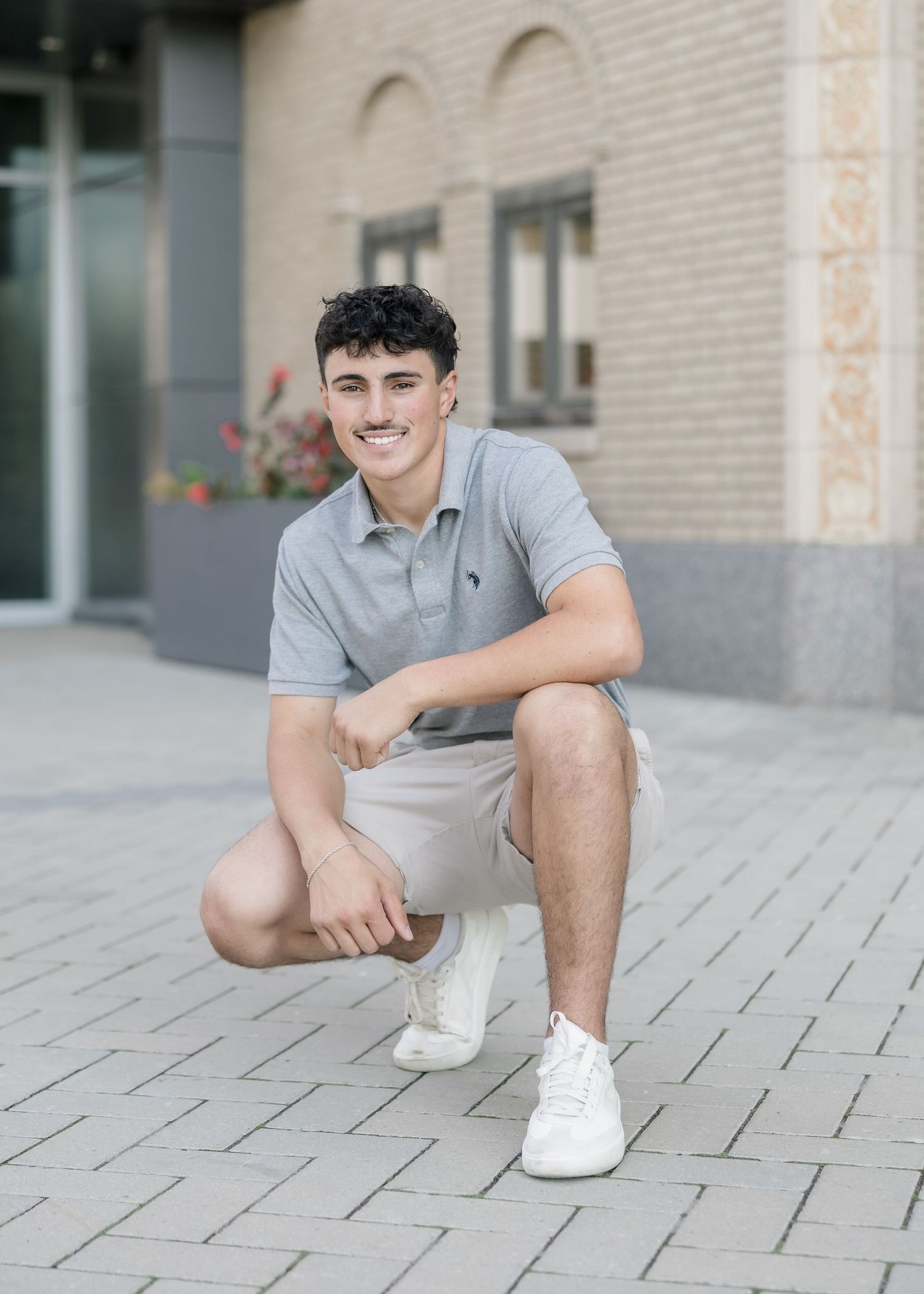 Person in a gray polo shirt and shorts sitting casually on stone pavement with white sneakers.