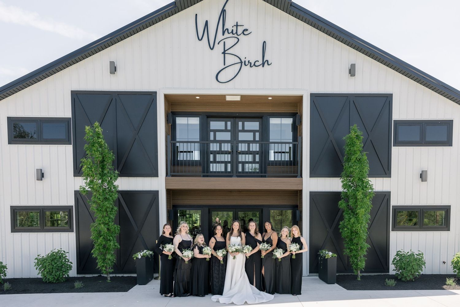 Bridal party in black dresses poses with bride in white gown outside White Birch barn wedding venue.