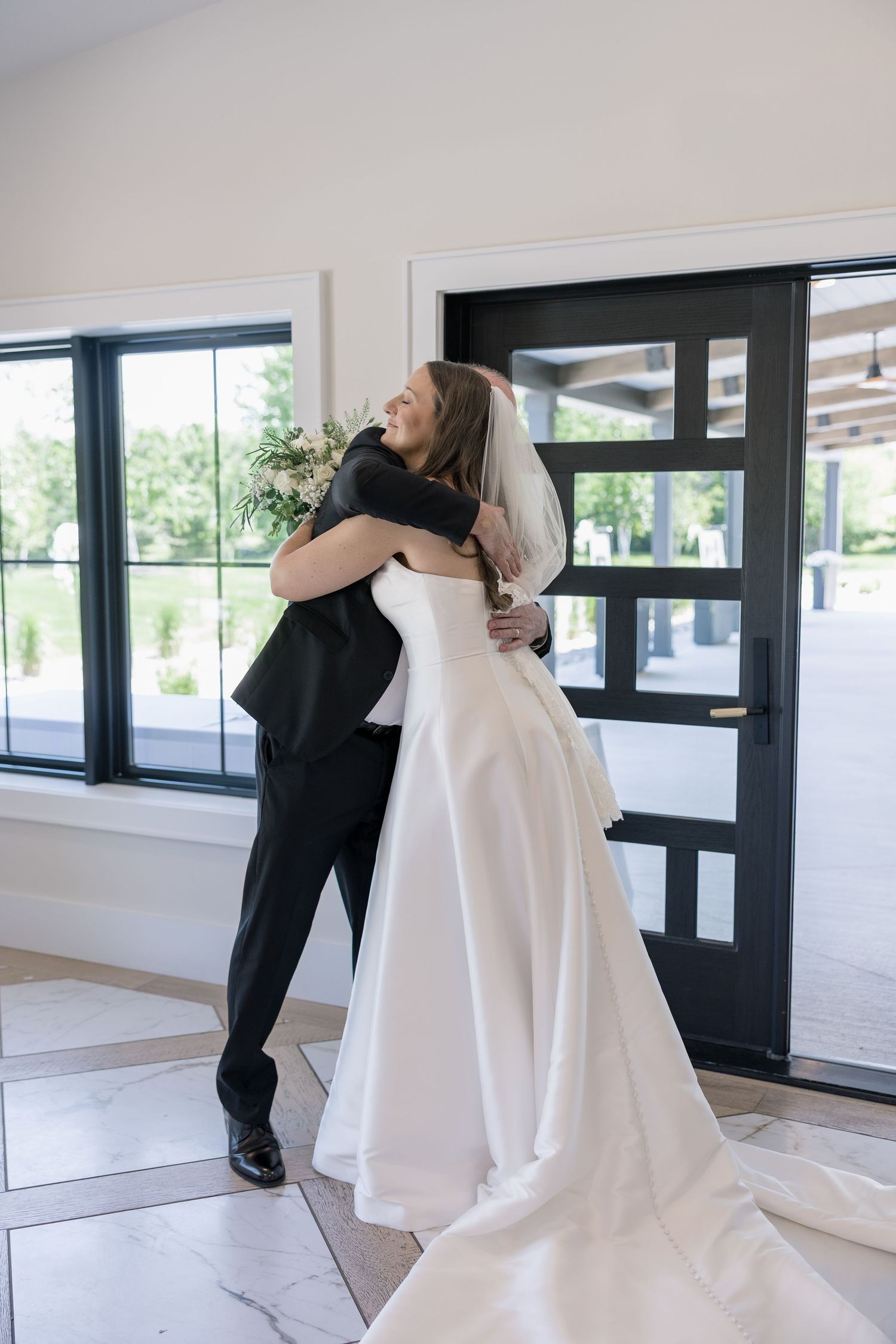 Bride and groom sharing an emotional first look hug inside bright venue with large windows.