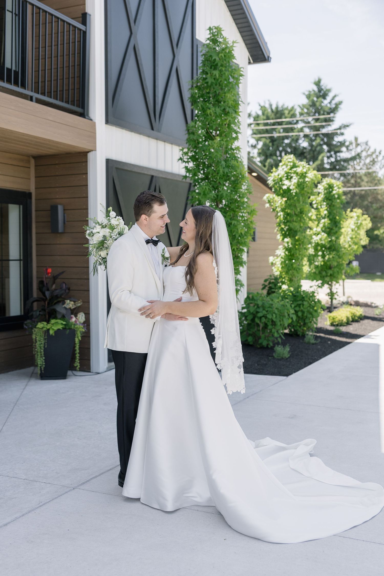A bride and groom share a romantic moment outdoors, with the bride in a flowing white gown and veil, groom in a white tuxedo.