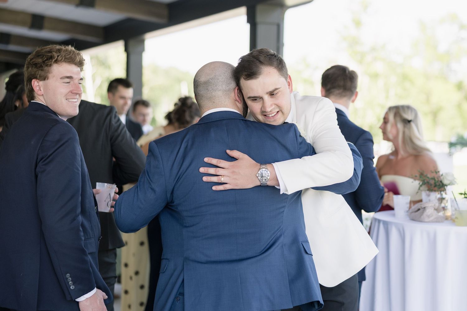 Two men in suits share a warm hug at a wedding reception while guests mingle in the background.
