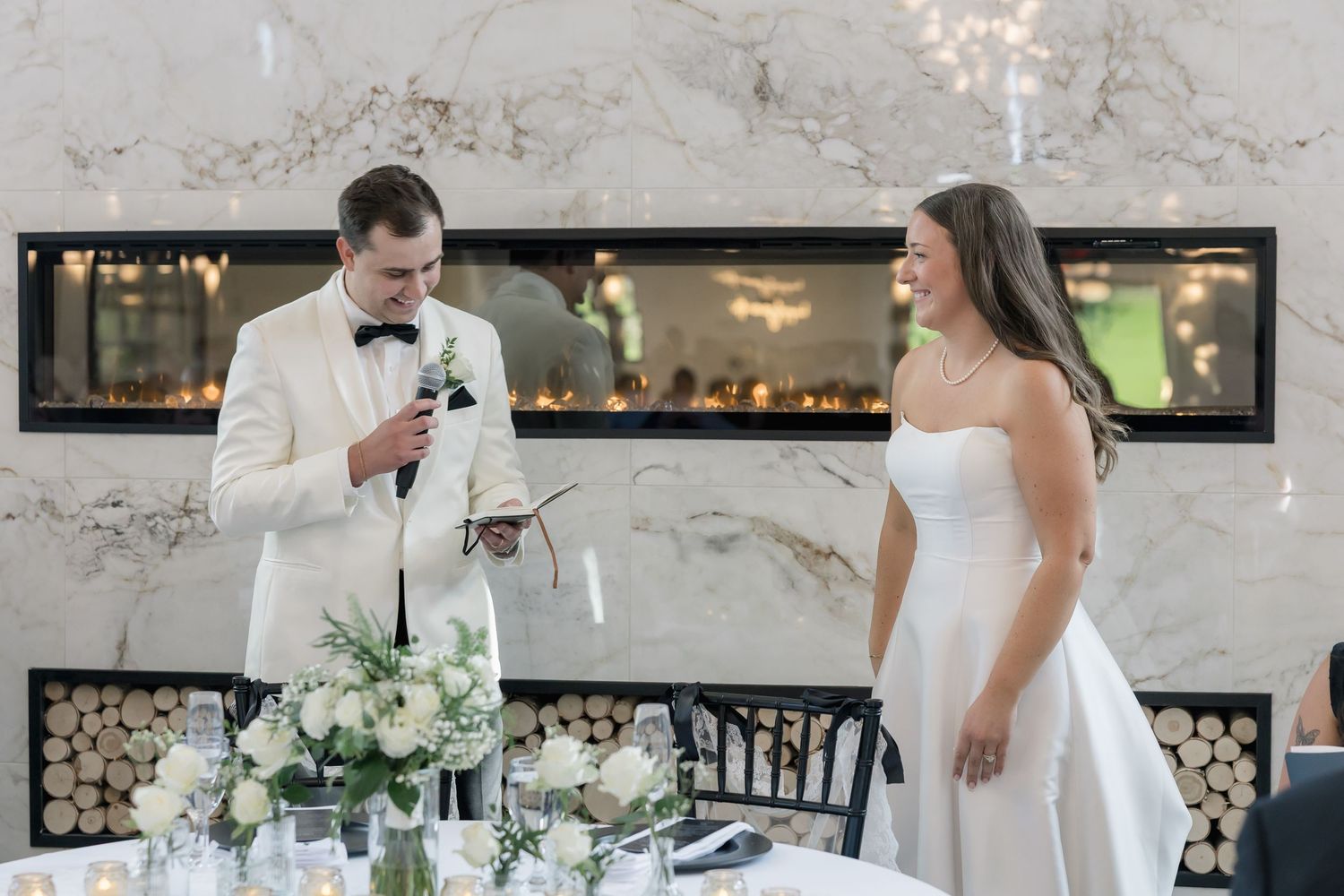 Groom in white tuxedo reads wedding speech while bride in strapless gown smiles at elegant marble reception venue.