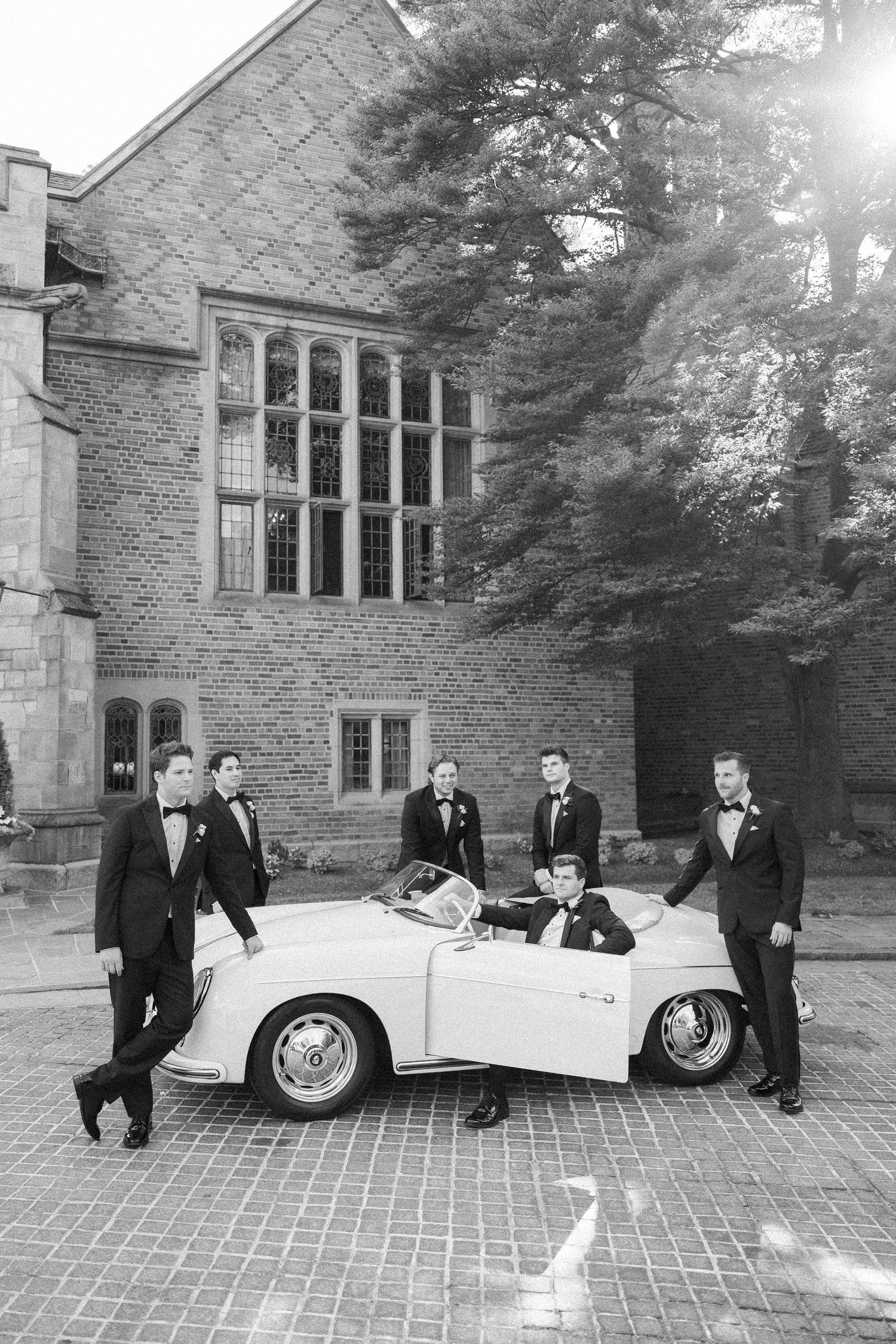 Vintage white car parked outside stone building with wedding party in formal attire.