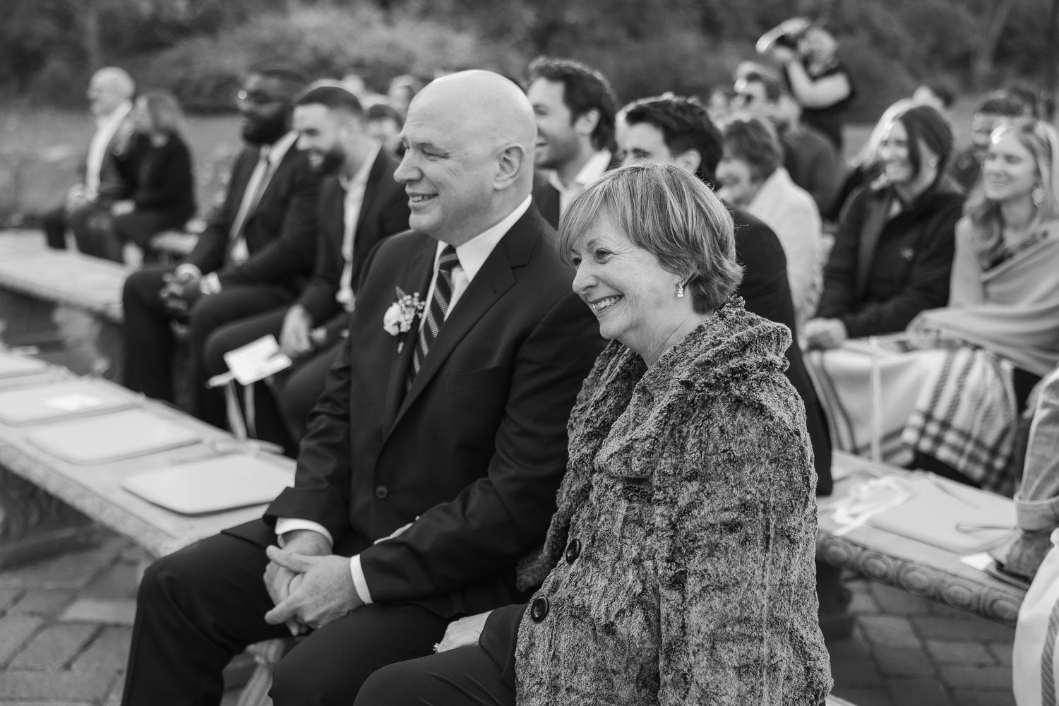 Wedding guests smile and enjoy outdoor ceremony from wooden benches.