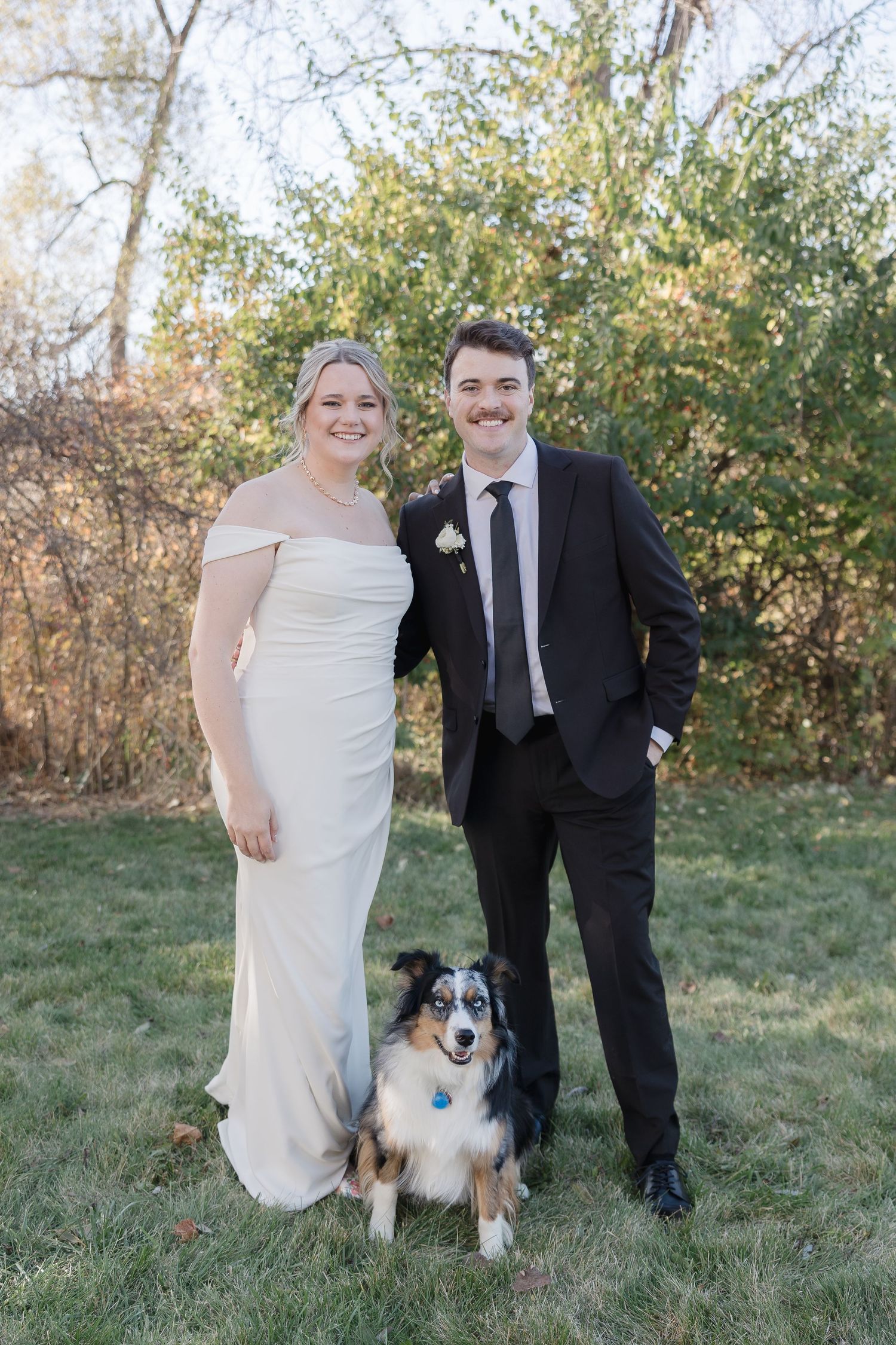 Bride and groom pose with their Australian Shepherd dog on grassy lawn during autumn wedding.