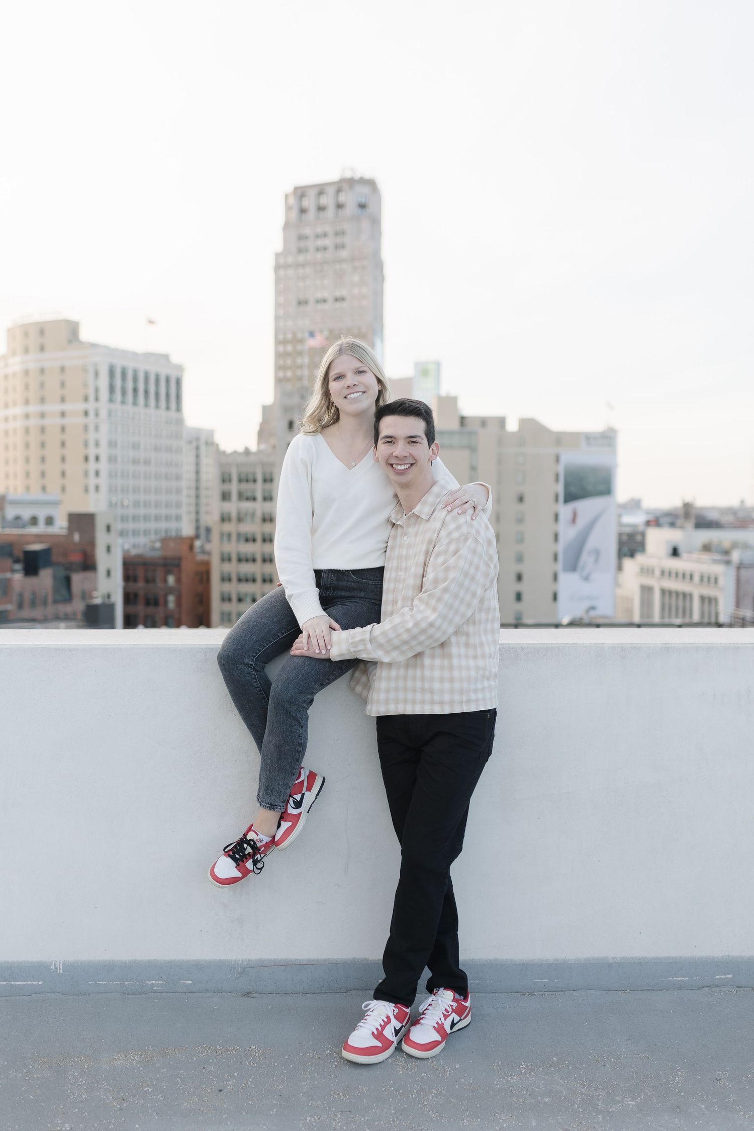 A happy couple in matching sweaters poses casually on a rooftop with urban architecture behind them.