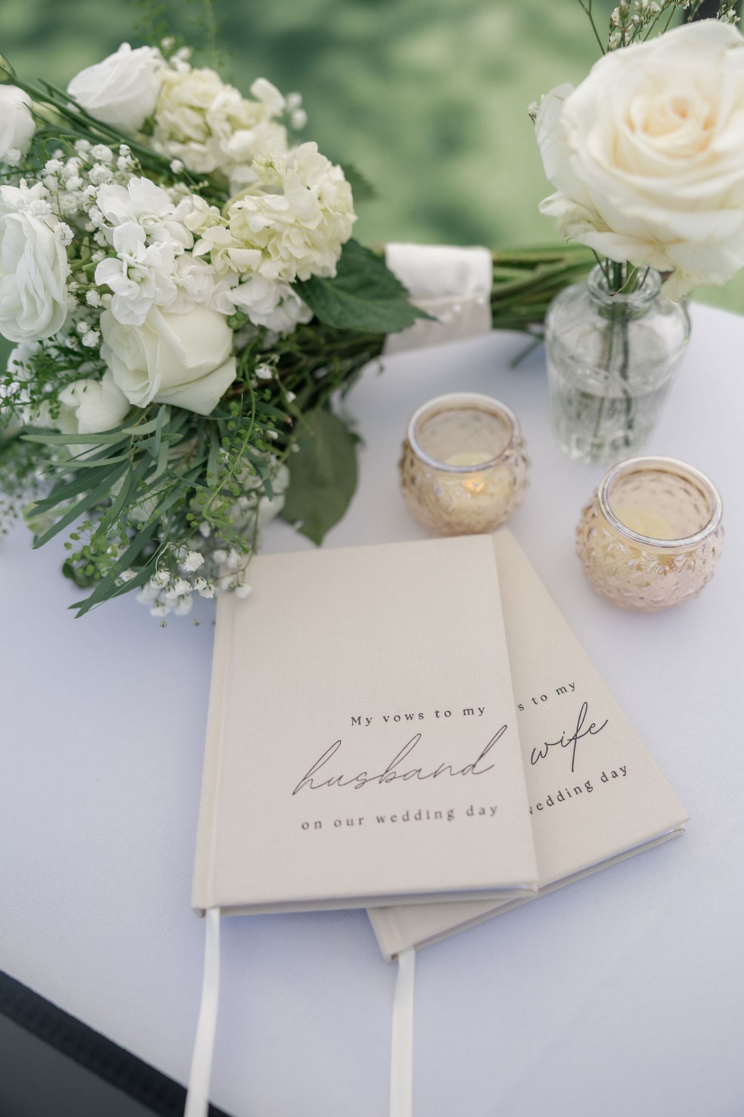 Wedding vow books for husband and wife on white table with white roses bouquet and gold tealight candles.
