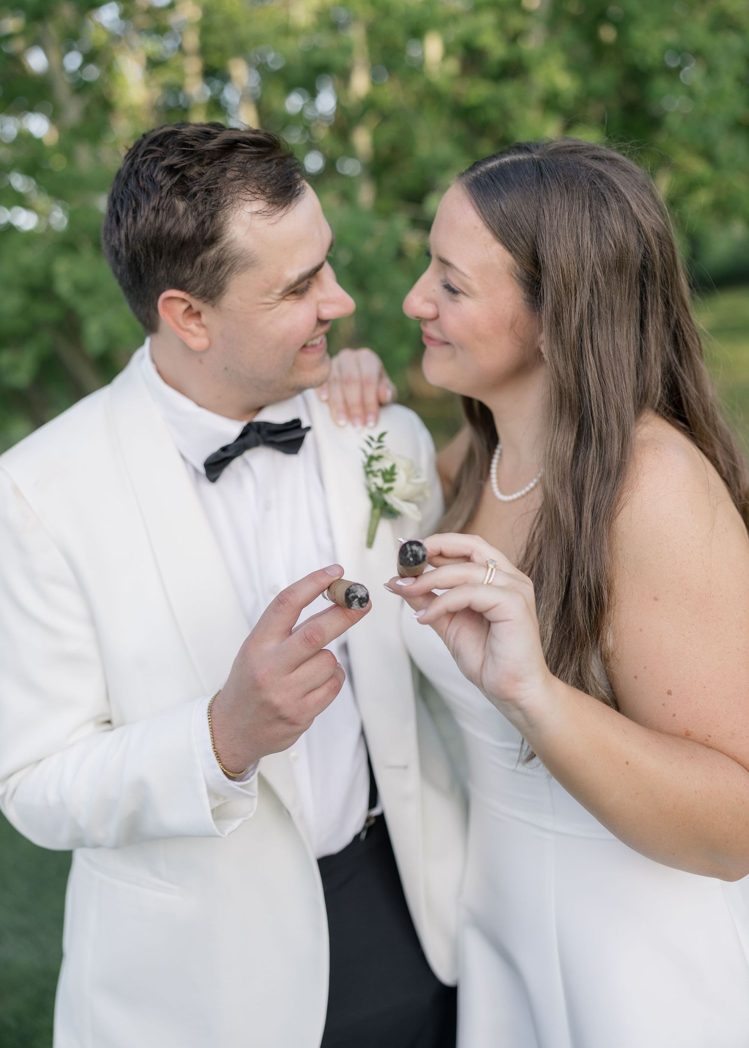 Bride and groom in elegant wedding attire smile at each other while holding small chocolates outdoors in a garden setting.