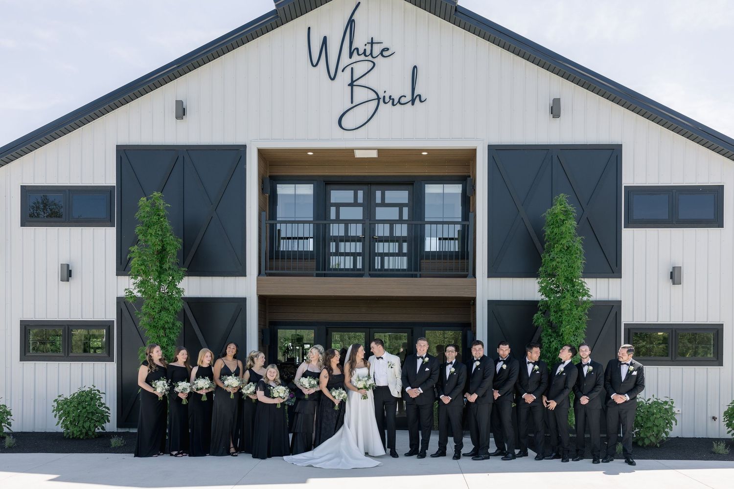 Wedding party poses in black attire before White Birch barn venue, bride in white gown centered.