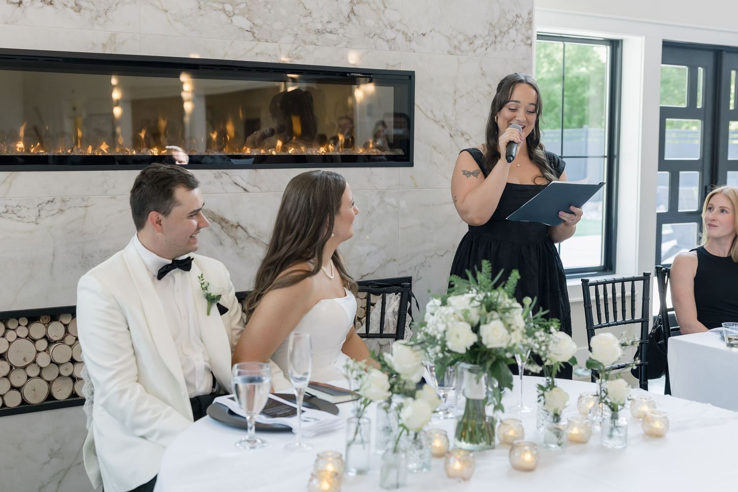 Bridesmaid delivers heartfelt speech at wedding reception beside marble fireplace and floral centerpieces.