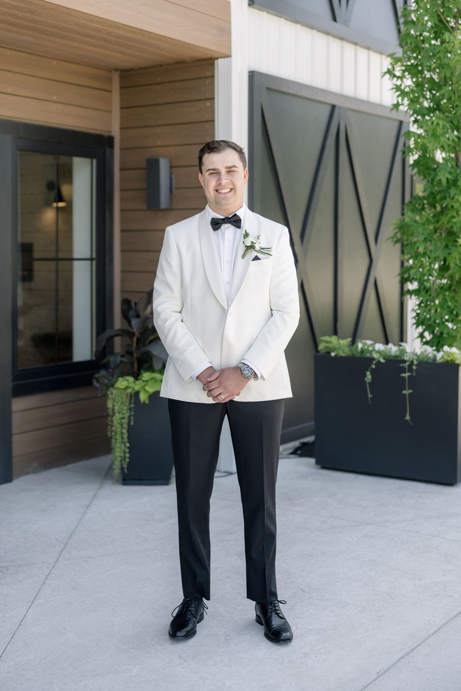 Groom in white tuxedo jacket and black bow tie smiles outdoors with boutonniere.