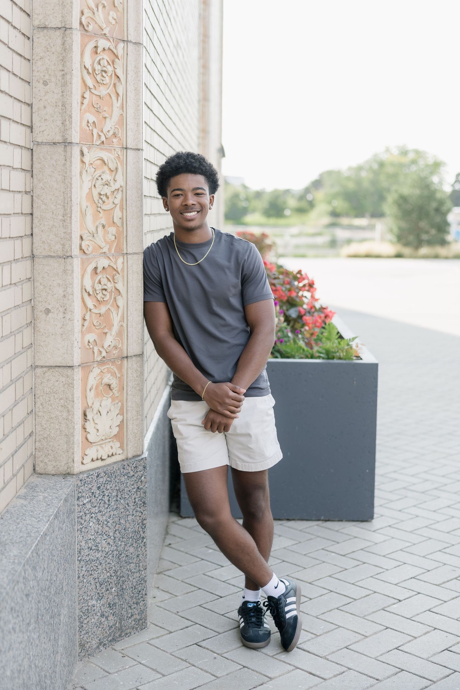 A person sits on a stone ledge wearing a grey t-shirt and shorts near decorative planters with flowers.