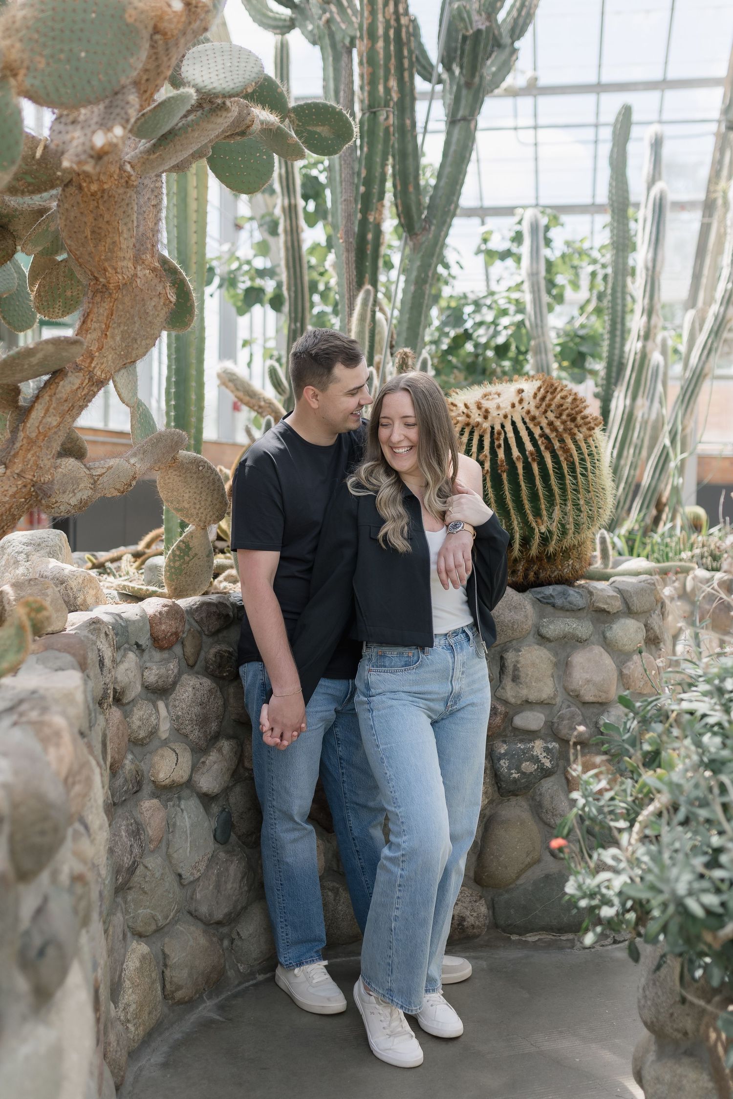 A couple stands together in a greenhouse surrounded by desert plants and cacti on a stone wall.