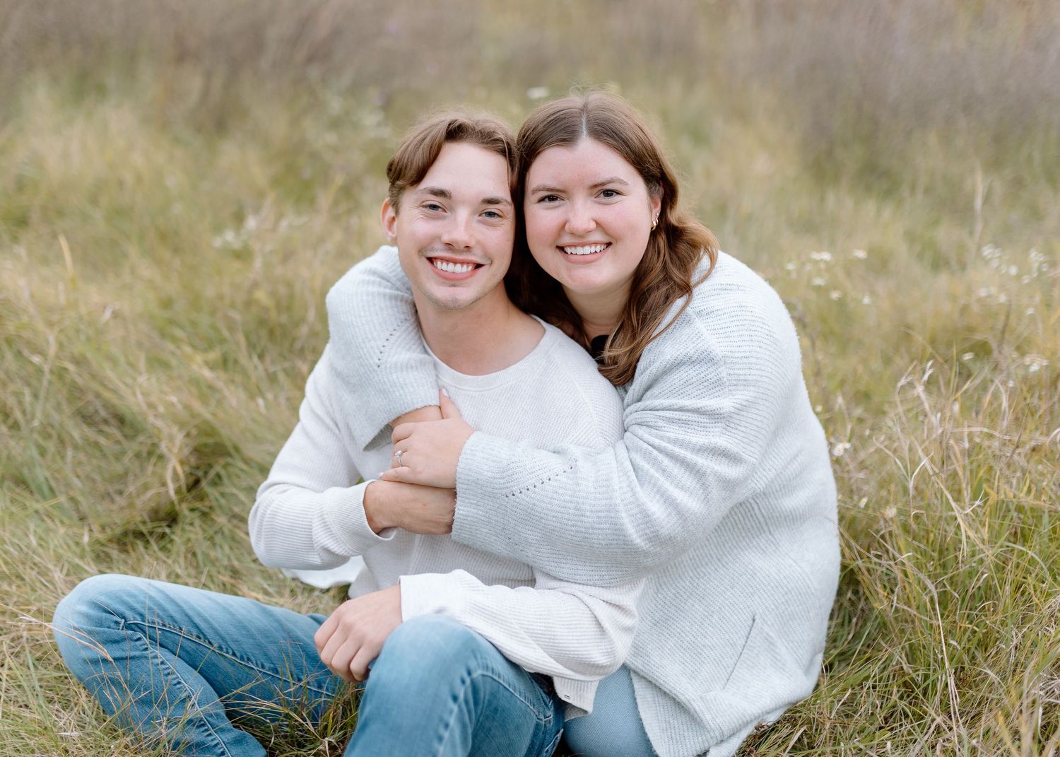 Two people in white sweaters sharing a joyful embrace while sitting outdoors in a grassy field.