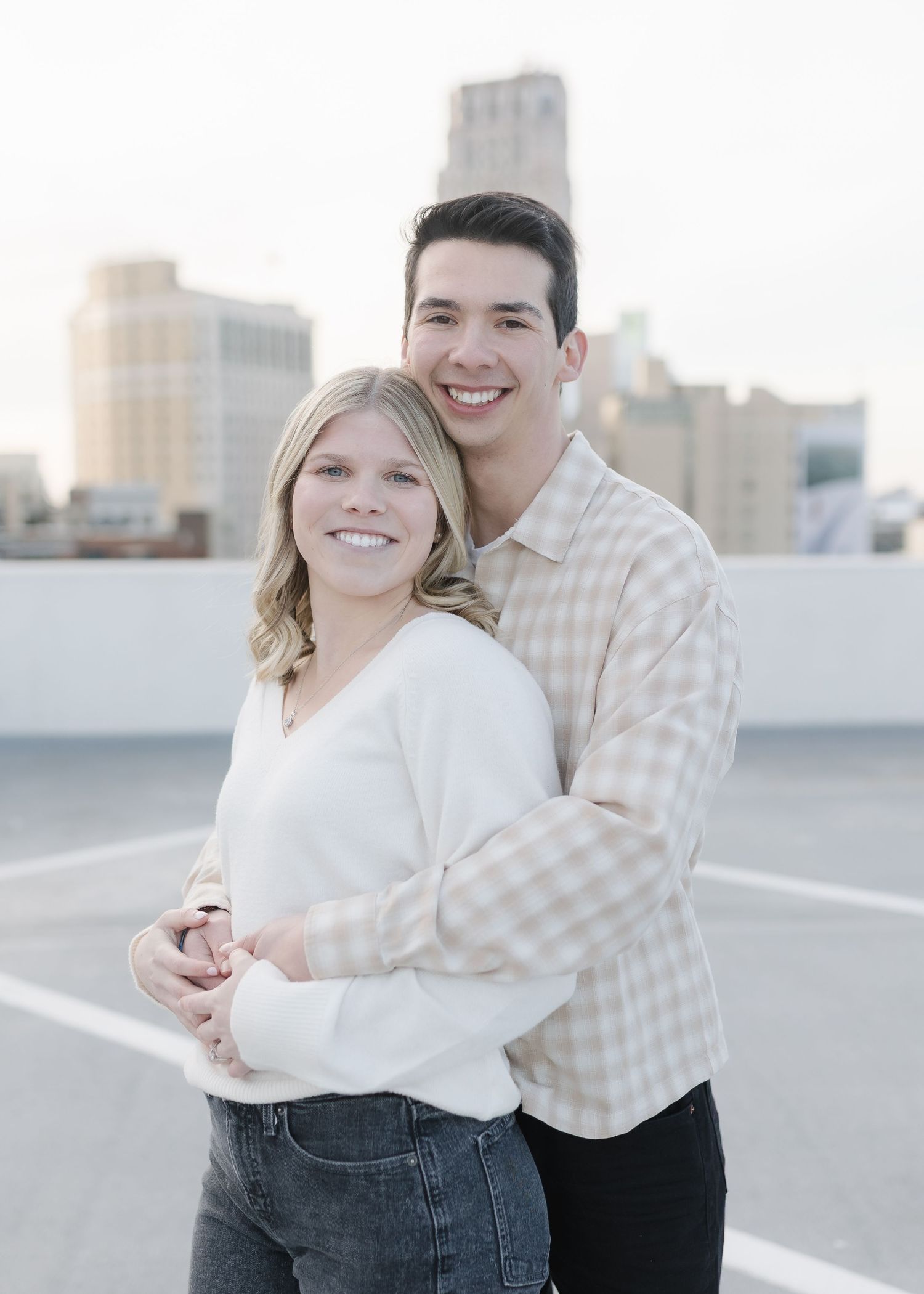A couple poses together on a rooftop with city buildings visible in the background during golden hour.